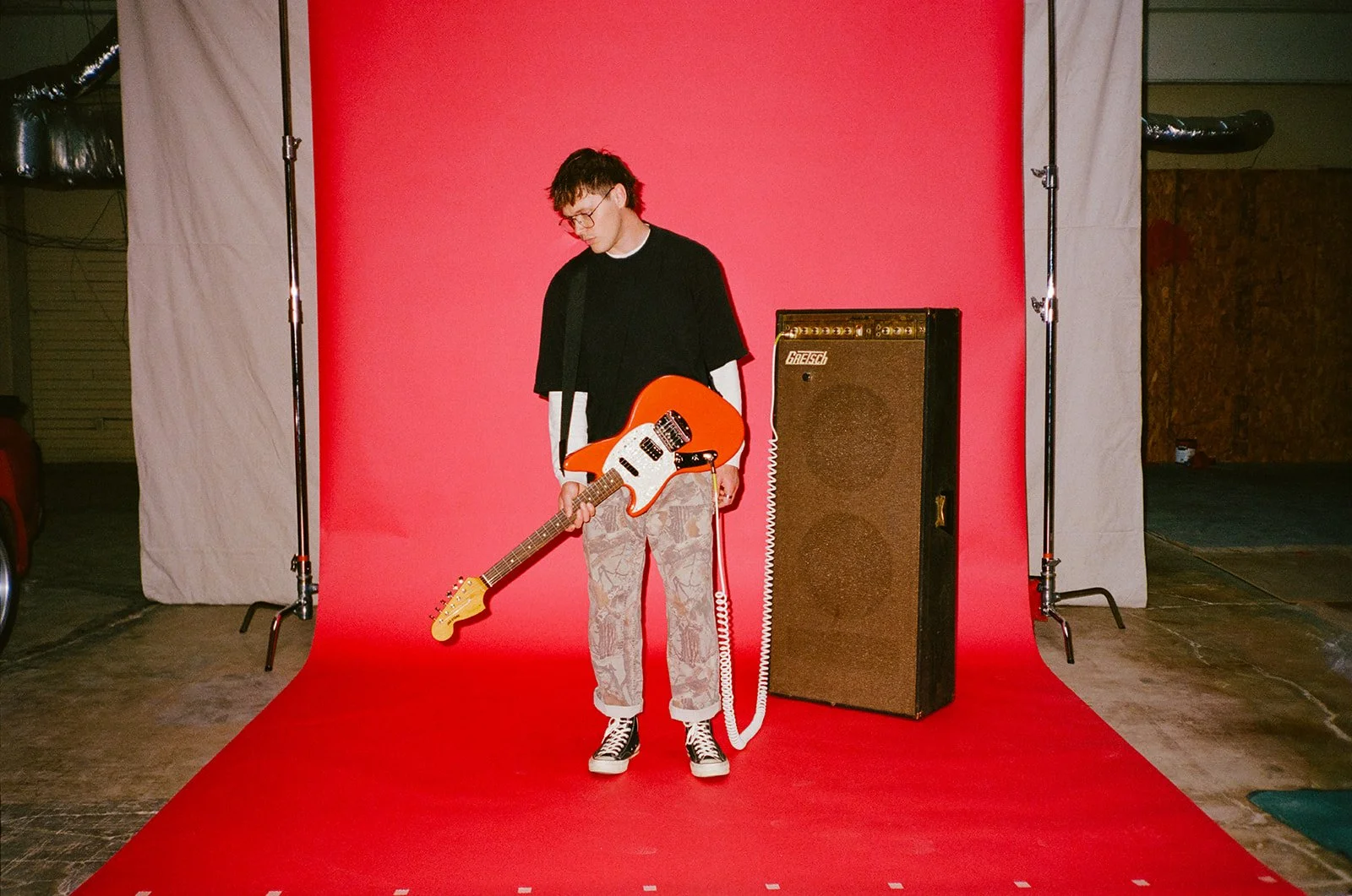 A young man with glasses and camouflage pants holding an orange electric guitar stands on a red photo backdrop, with an amplifier nearby.