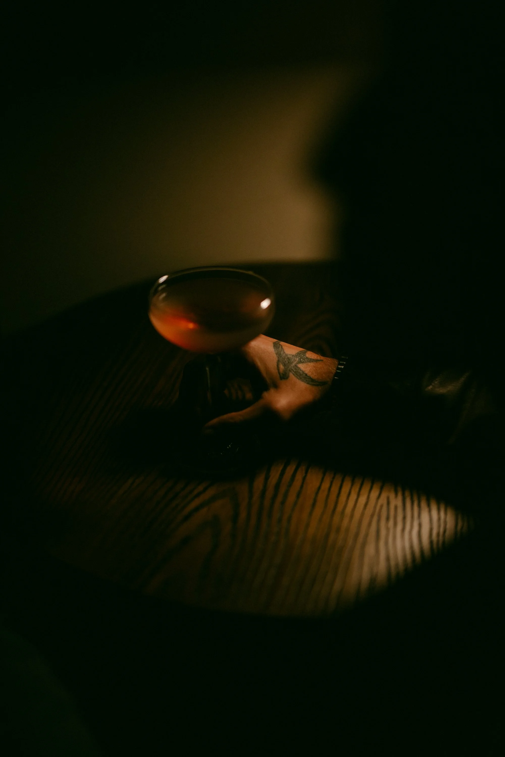A glass of wine on a table with striped fabric, dark background, and a tattooed arm nearby.