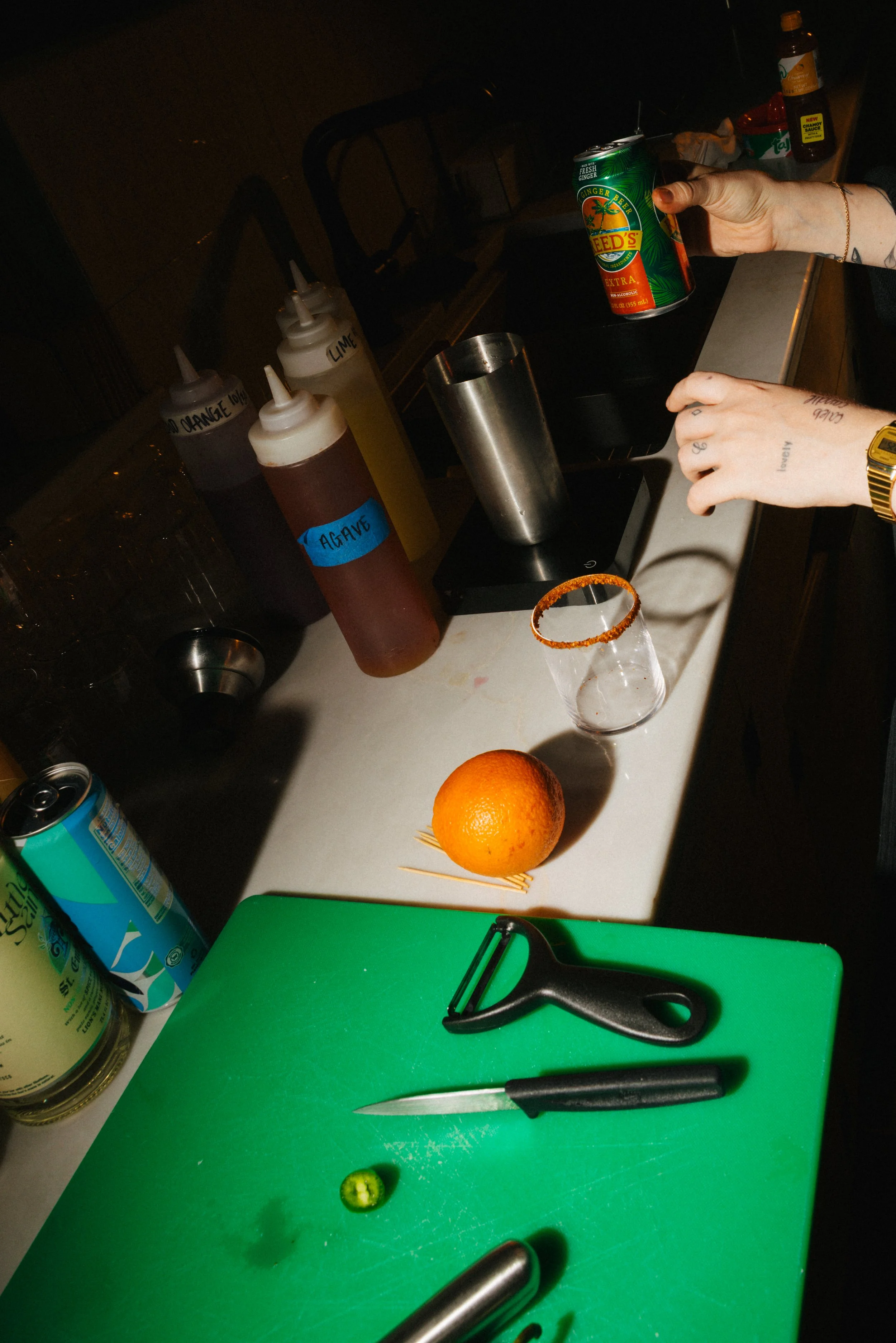 Bar countertop with orange, lime, cocktail tools, a can of soda, a glass rimmed with chili powder, and drink ingredients including juices and garnishes.
