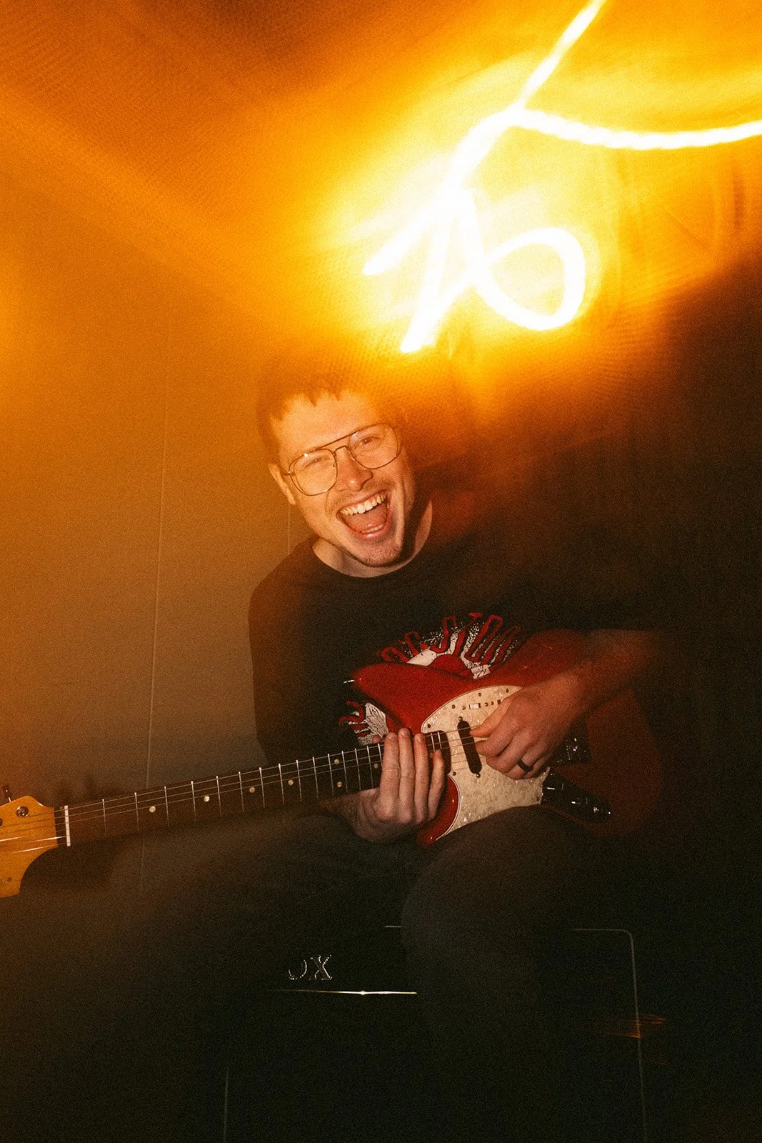 Joyful man with glasses playing an electric guitar in a dim room with bright orange lighting and neon sign in the background.