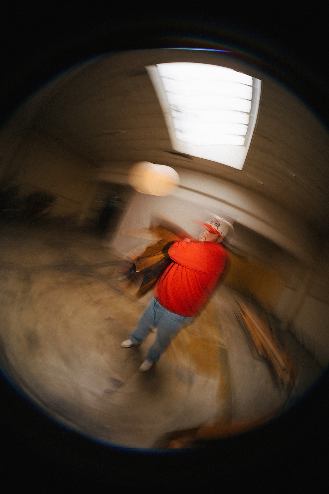 A person wearing a red shirt, blue jeans, and a white cap with a red logo in an indoor setting, taken with a fisheye lens, creating a circular, distorted view.