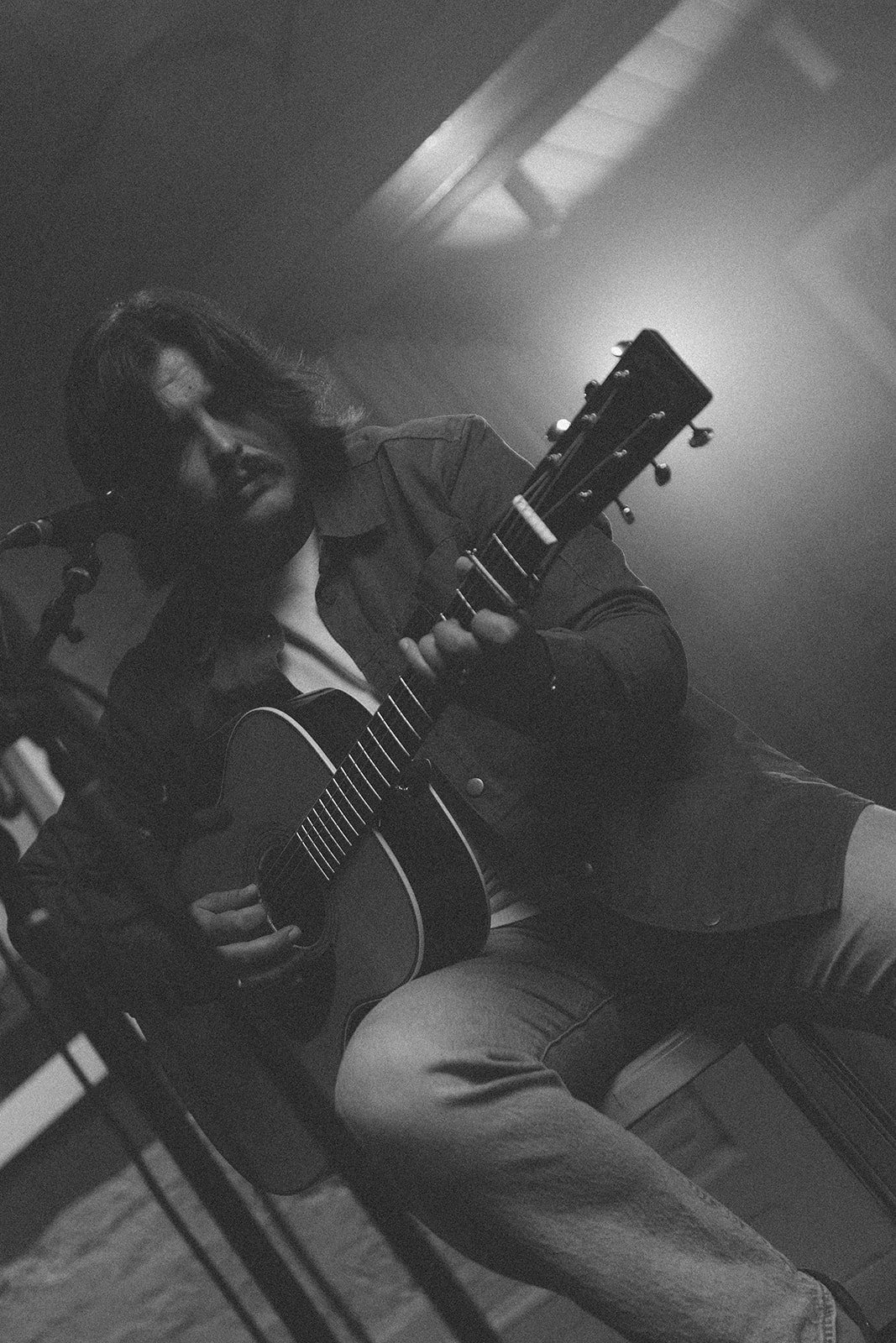 A woman with shoulder-length hair, wearing a dark jacket, playing an acoustic guitar in a dimly lit room with a microphone nearby.