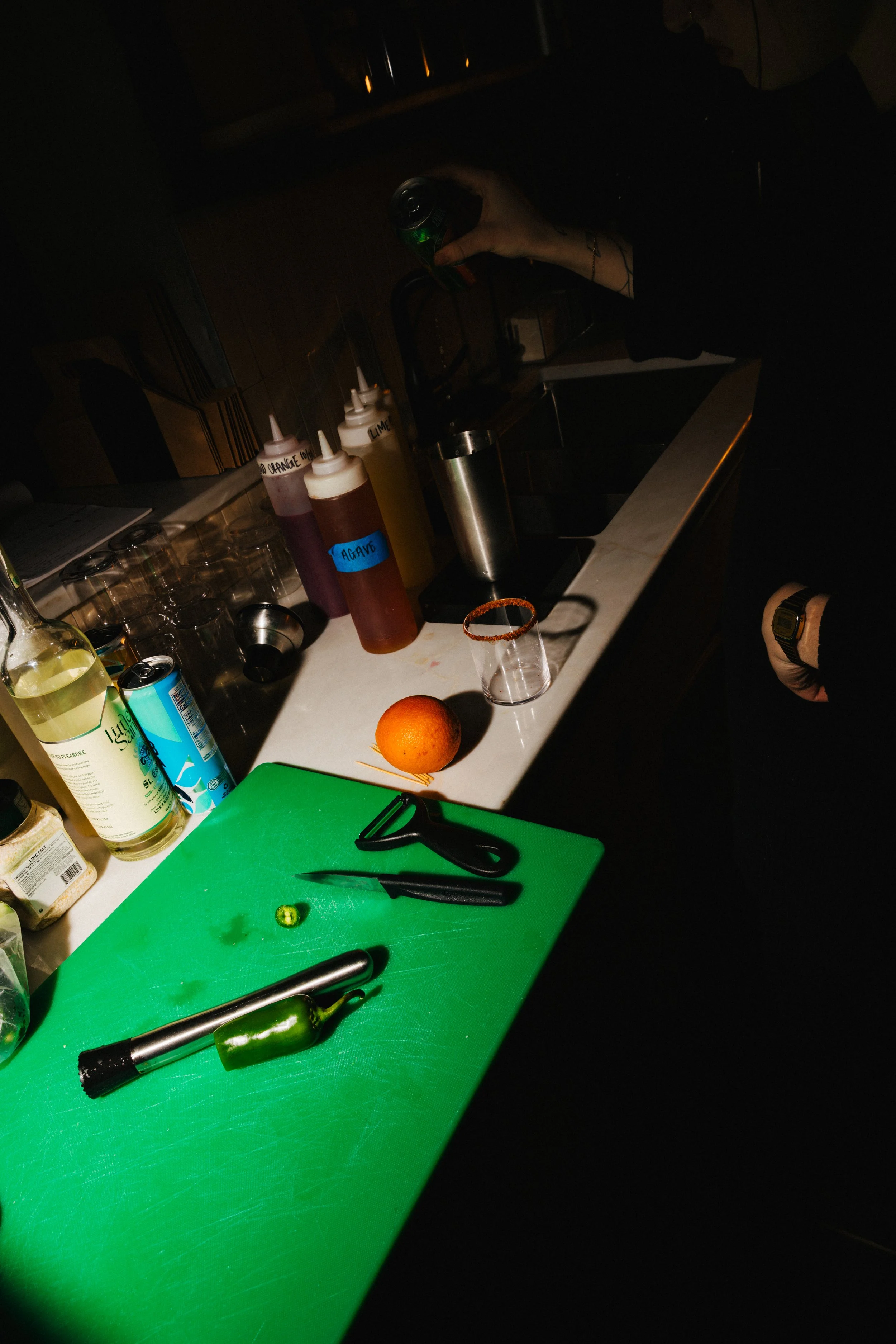 A bartender's workspace with various bottles of juices, a lime, a knife, a peeler, and a green cutting board on a white countertop. A person is pouring a drink from a can, with bottles labeled in the background.