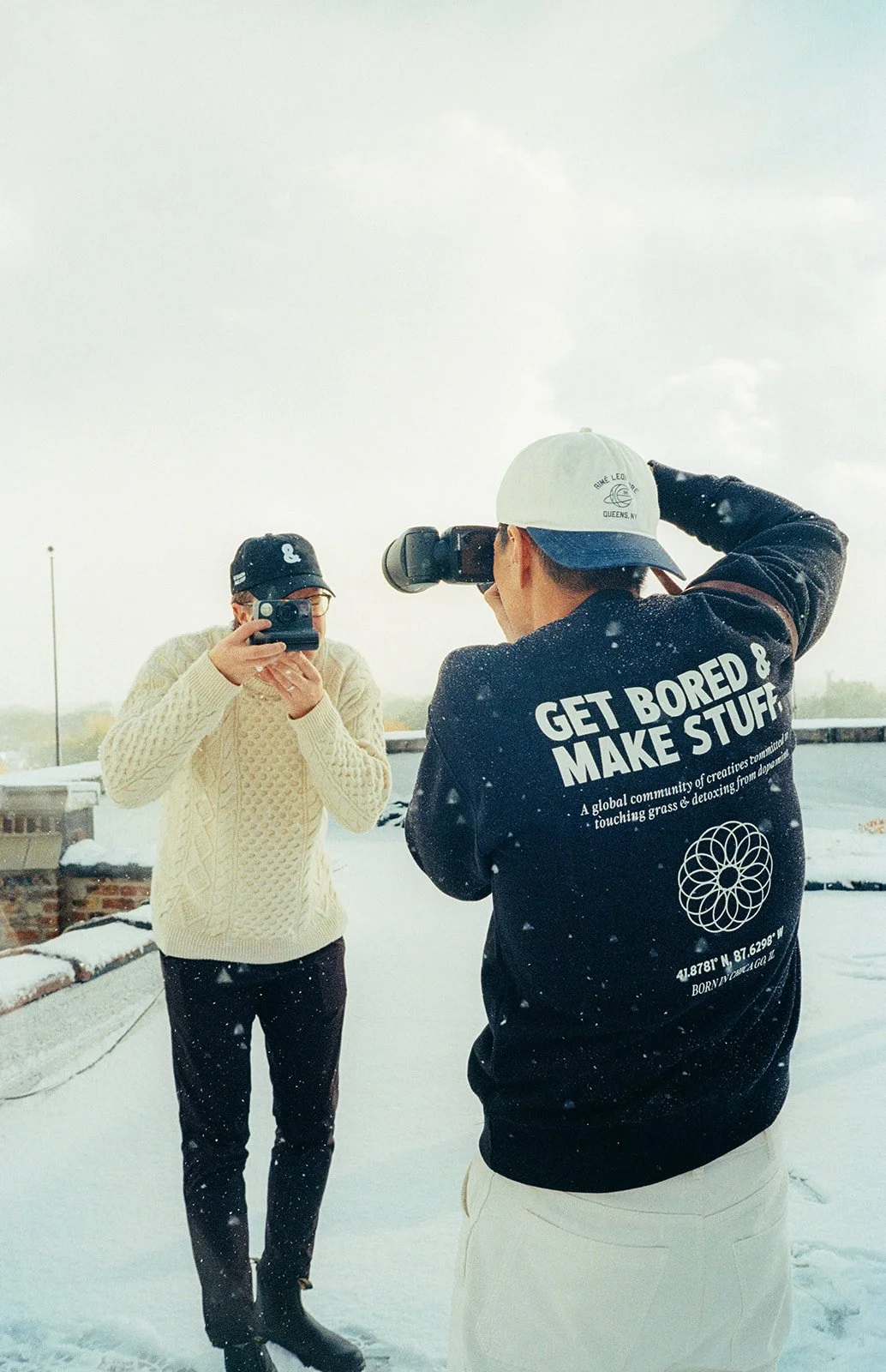 Two people standing outdoors in the snow, one taking a photo or video with a phone, the other holding a camera. The person holding the camera is wearing a black jacket with white text on the back that says 'GET BORED & MAKE STUFF,' and a black aviato