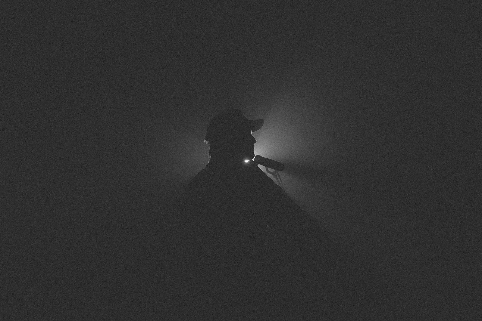 Silhouette of a person wearing a hat, facing left, with a flashlight on their neck in a dark, foggy environment.