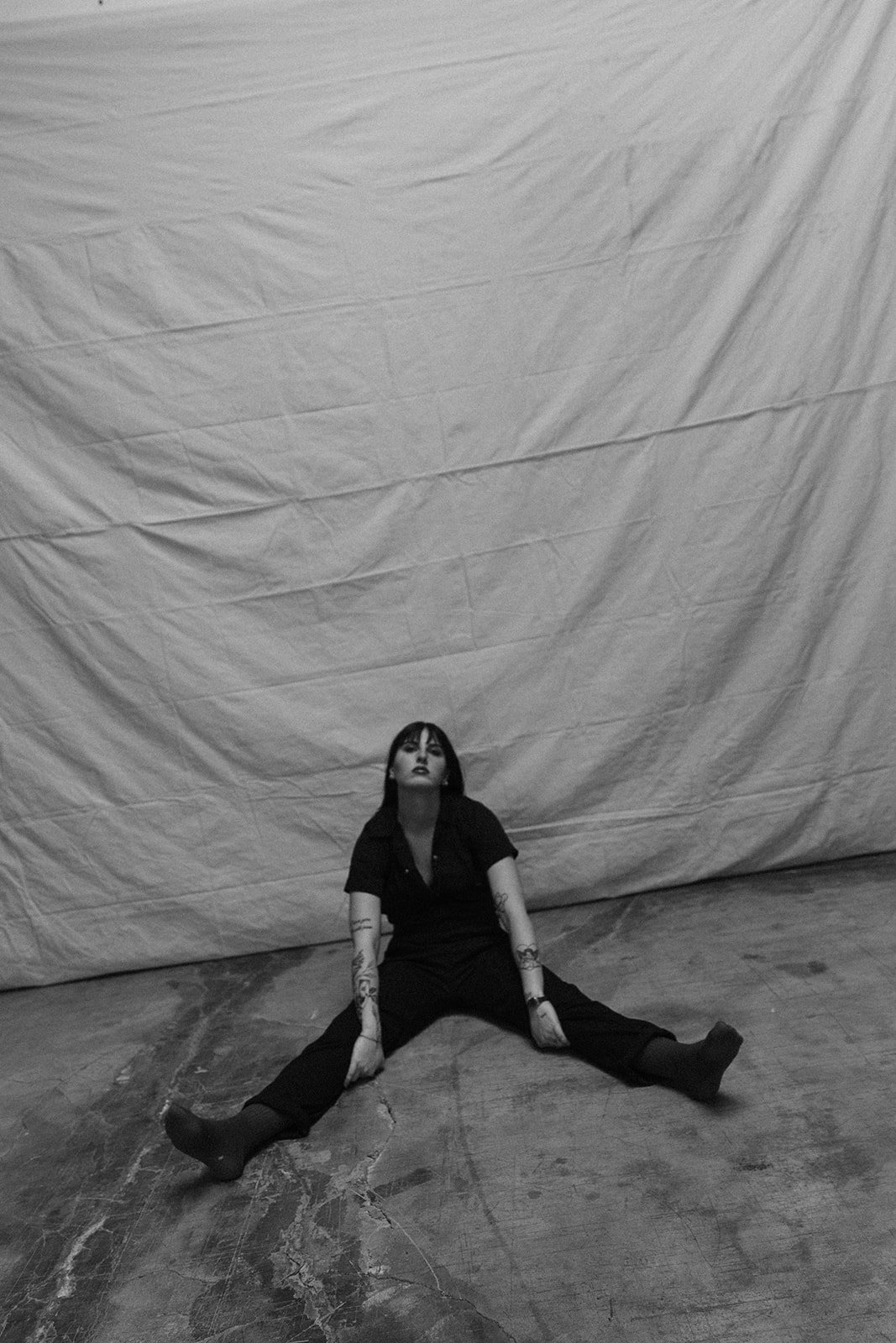 A woman with dark hair and tattoos, dressed in black, sitting on the floor with legs spread apart, in front of a large, wrinkled backdrop.