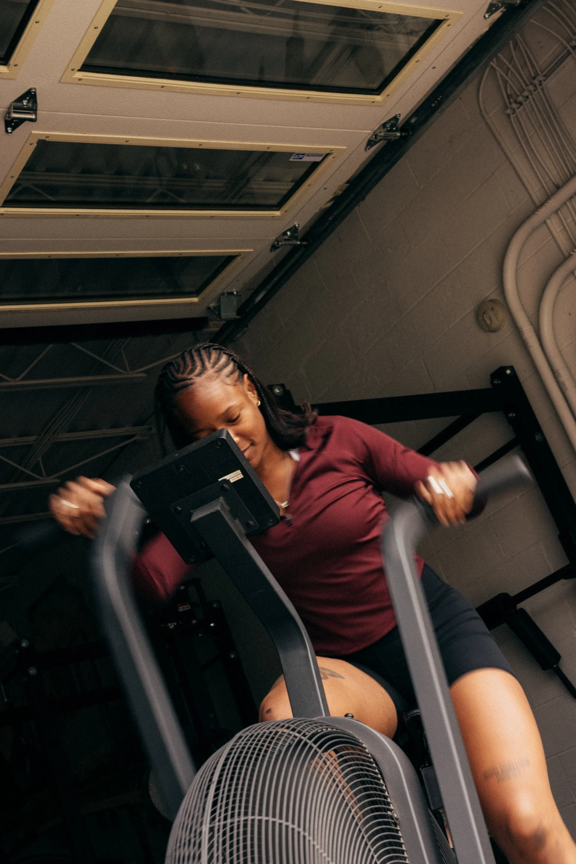 A woman with braided hair wearing a maroon shirt and black shorts, exercising on an elliptical machine in a gym with a large fan in the foreground and a wall with visible pipes in the background.