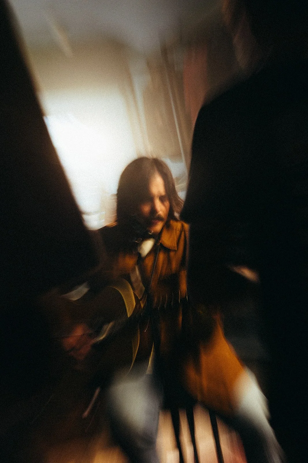 A man with long hair and a mustache playing an acoustic guitar, surrounded by blurred figures.