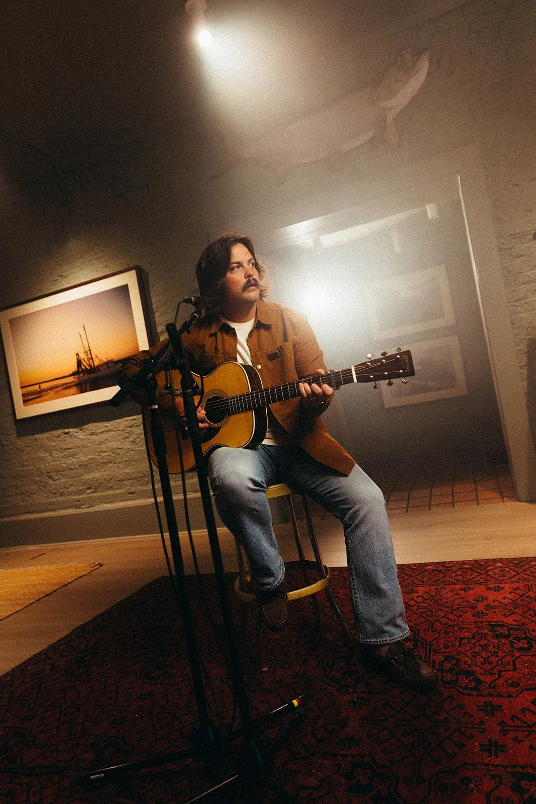 A man with shoulder-length hair and a mustache playing an acoustic guitar in a room with artsy decor, including framed art and a red patterned rug.
