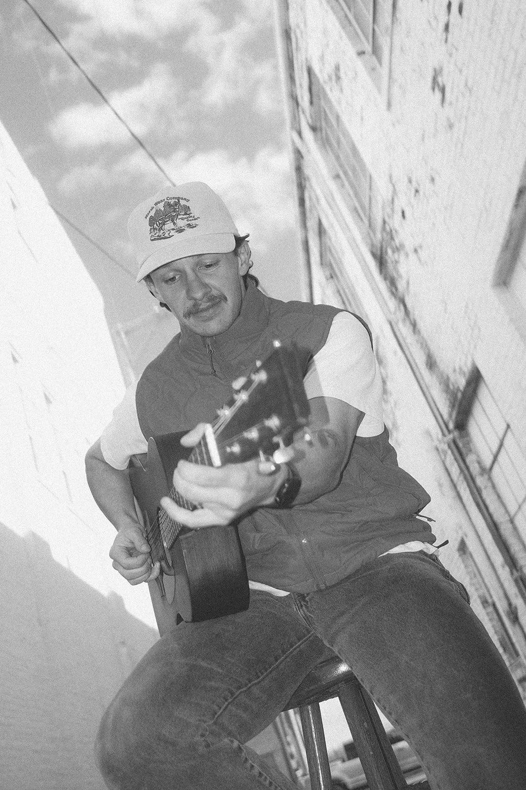 A man playing an acoustic guitar while seated on a chair outdoors on a city street, wearing a baseball cap and casual clothing.
