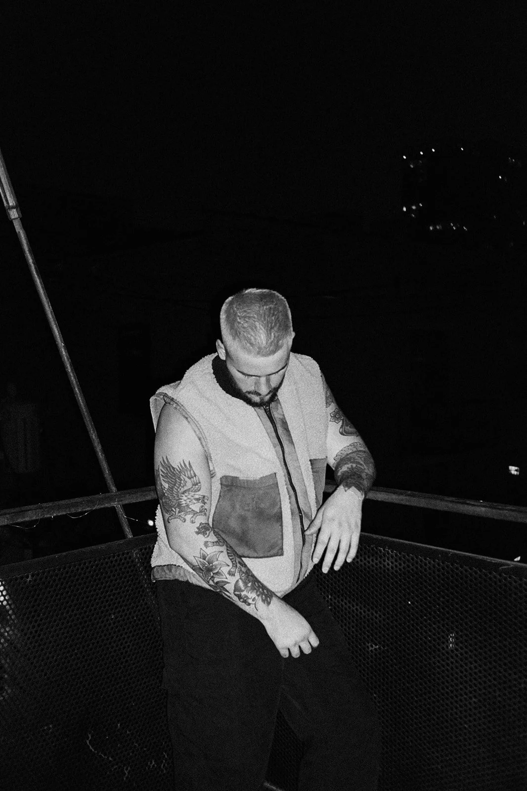 A tattooed man with a beard, wearing a sleeveless vest, stands on a rooftop at night, looking down with his hand on his waistband.