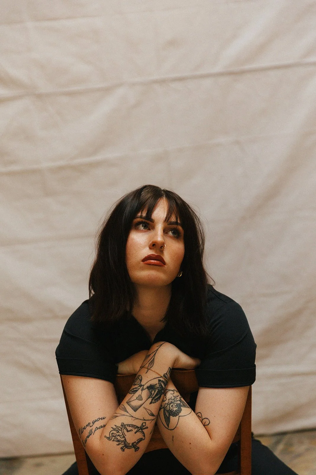 A woman with dark hair and tattoos on her arms, wearing a black shirt, sitting on a chair with her hands clasped under her chin, looking upward.