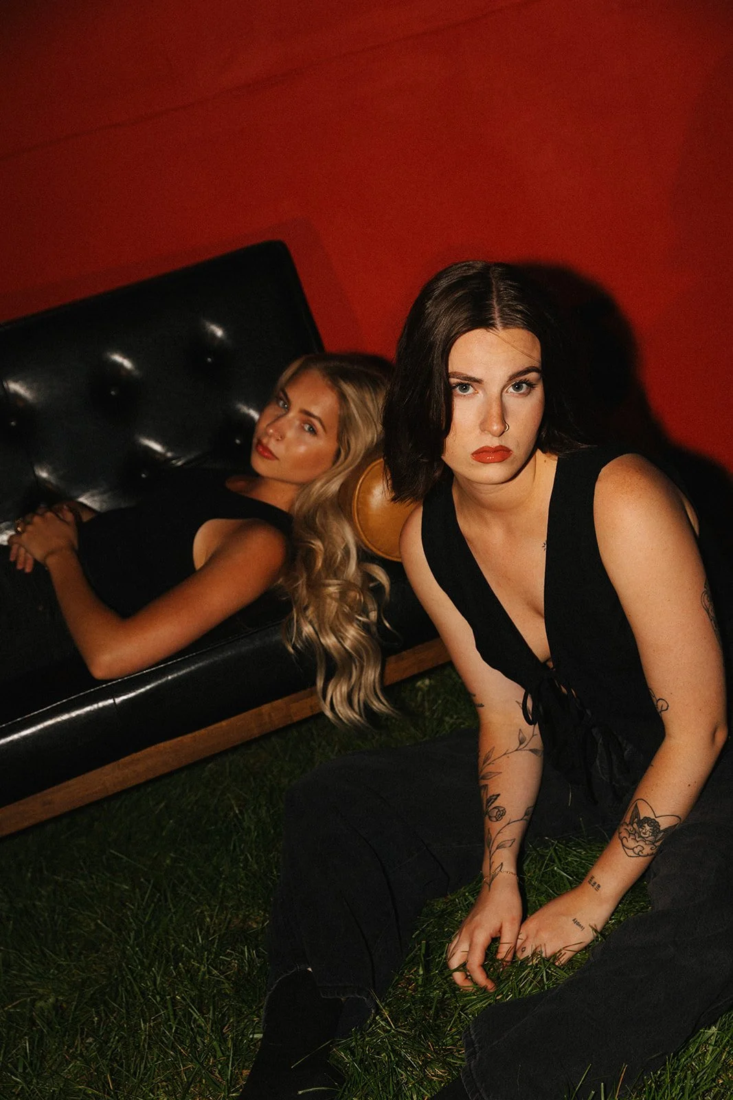 Two women, one with long blonde hair lying on a black leather couch, and the other with short dark hair sitting on grass in front of the couch, both wearing black outfits. The background is red.