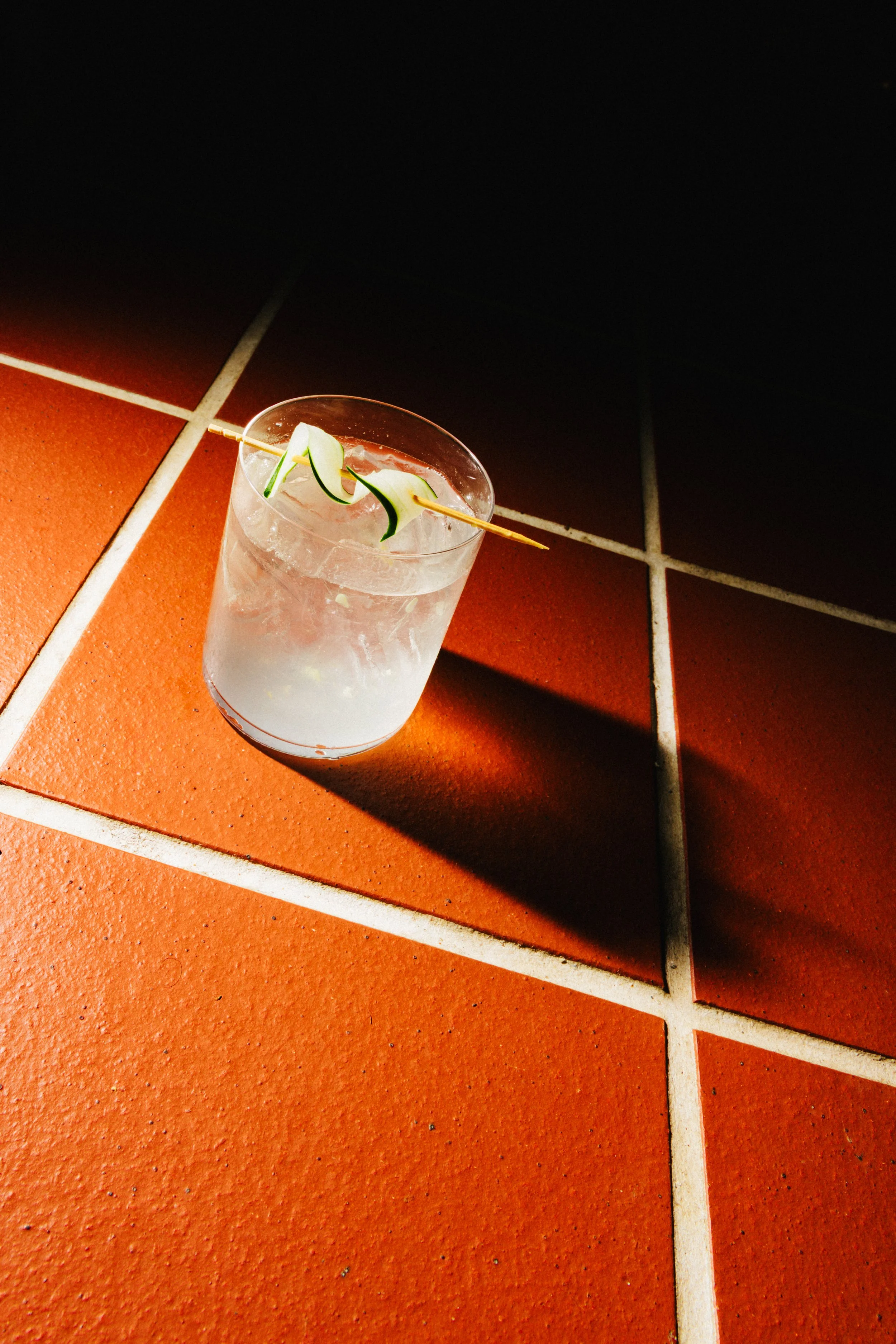 A clear glass containing a beverage with ice and thinly sliced cucumber, placed on a reddish-orange tiled floor, casting a shadow.