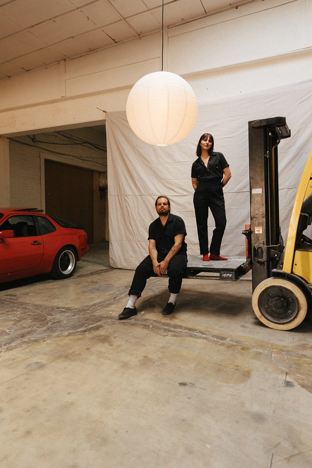 A man and a woman in black clothes posing in an empty industrial space with a red car, a large paper lantern, and a forklift. The woman is standing on a platform, and the man is sitting on the platform's edge.