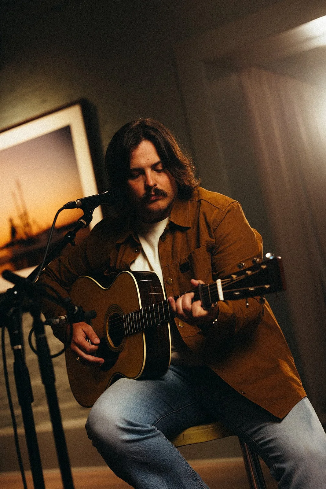 A man with shoulder-length dark hair and a mustache playing an acoustic guitar in a room with dim lighting, a microphone stand nearby, and a framed sunset photograph on the wall.