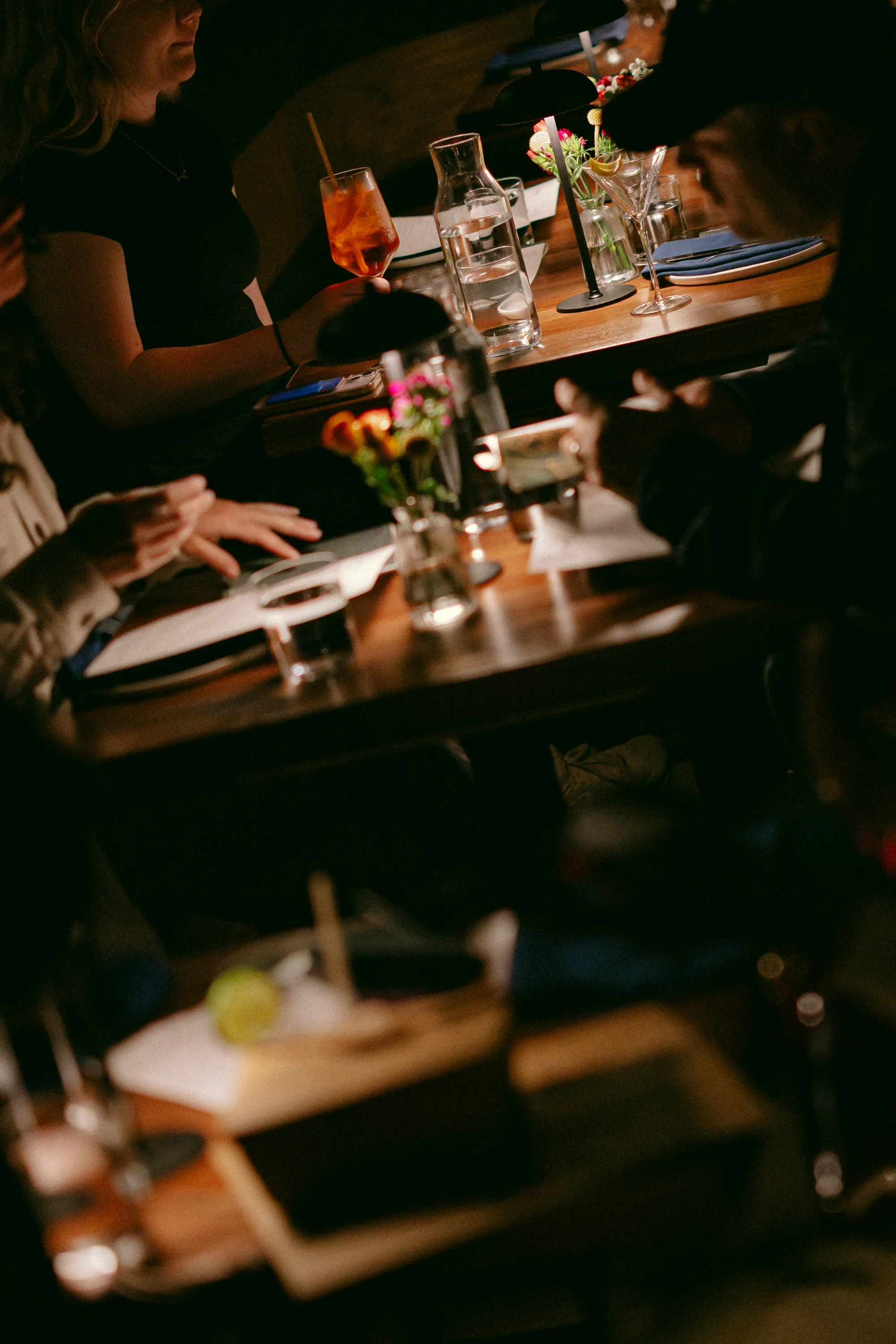 People sitting around a wooden table in a dimly lit restaurant or bar, with various drinks, flowers, and plates on the table.