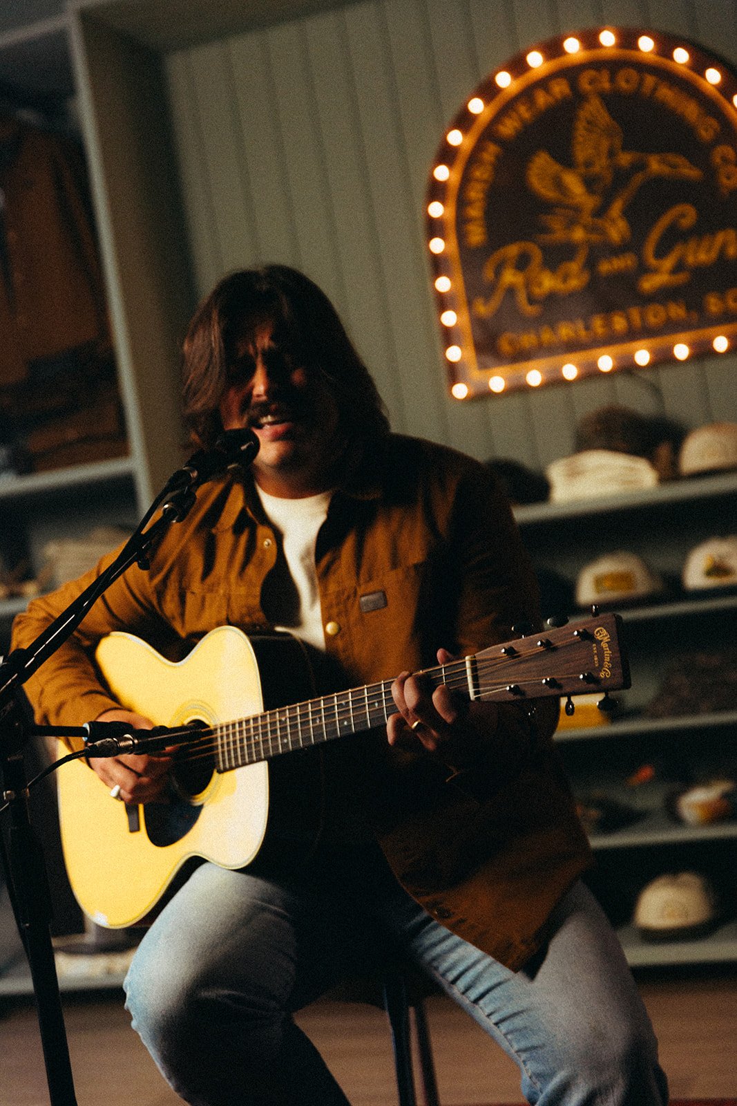 A person playing an acoustic guitar and singing into a microphone during a live performance in a cozy, dimly lit setting, with a sign on the wall reading 'Since 1992' and a shelf with hats in the background.
