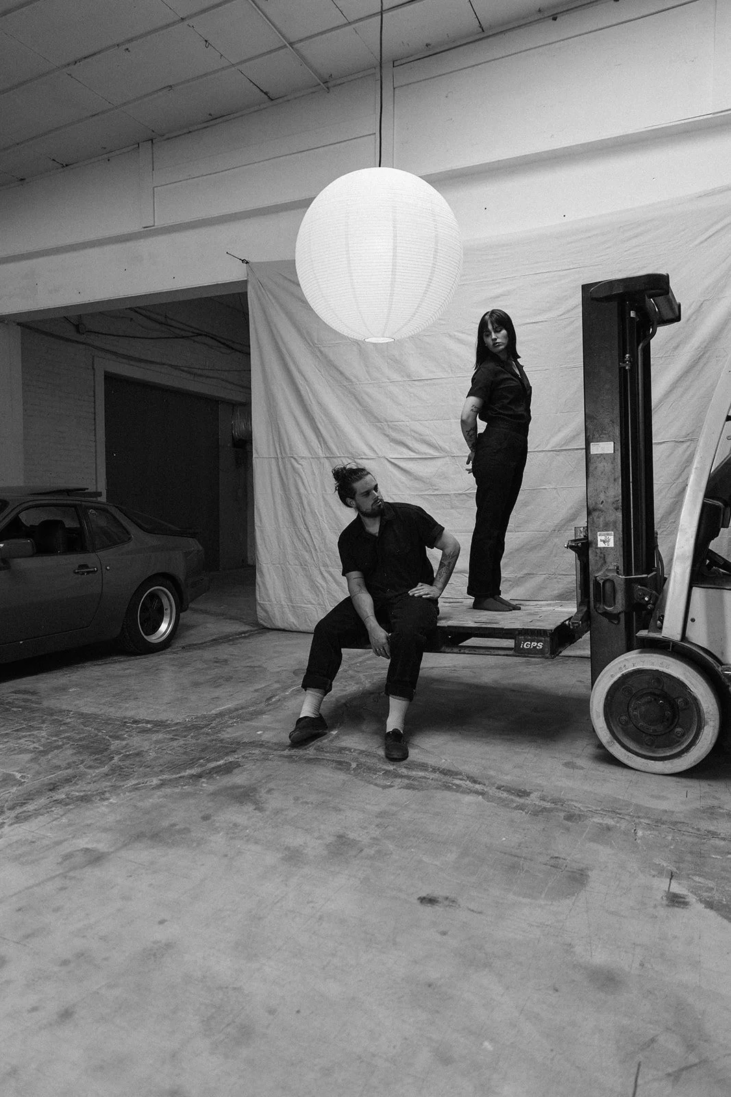 Black and white photo of two people in a warehouse or studio, with a vintage car on the left, a large paper lantern overhead, and a backdrop. One person is sitting on a forklift, and the other is standing on the forklift platform.