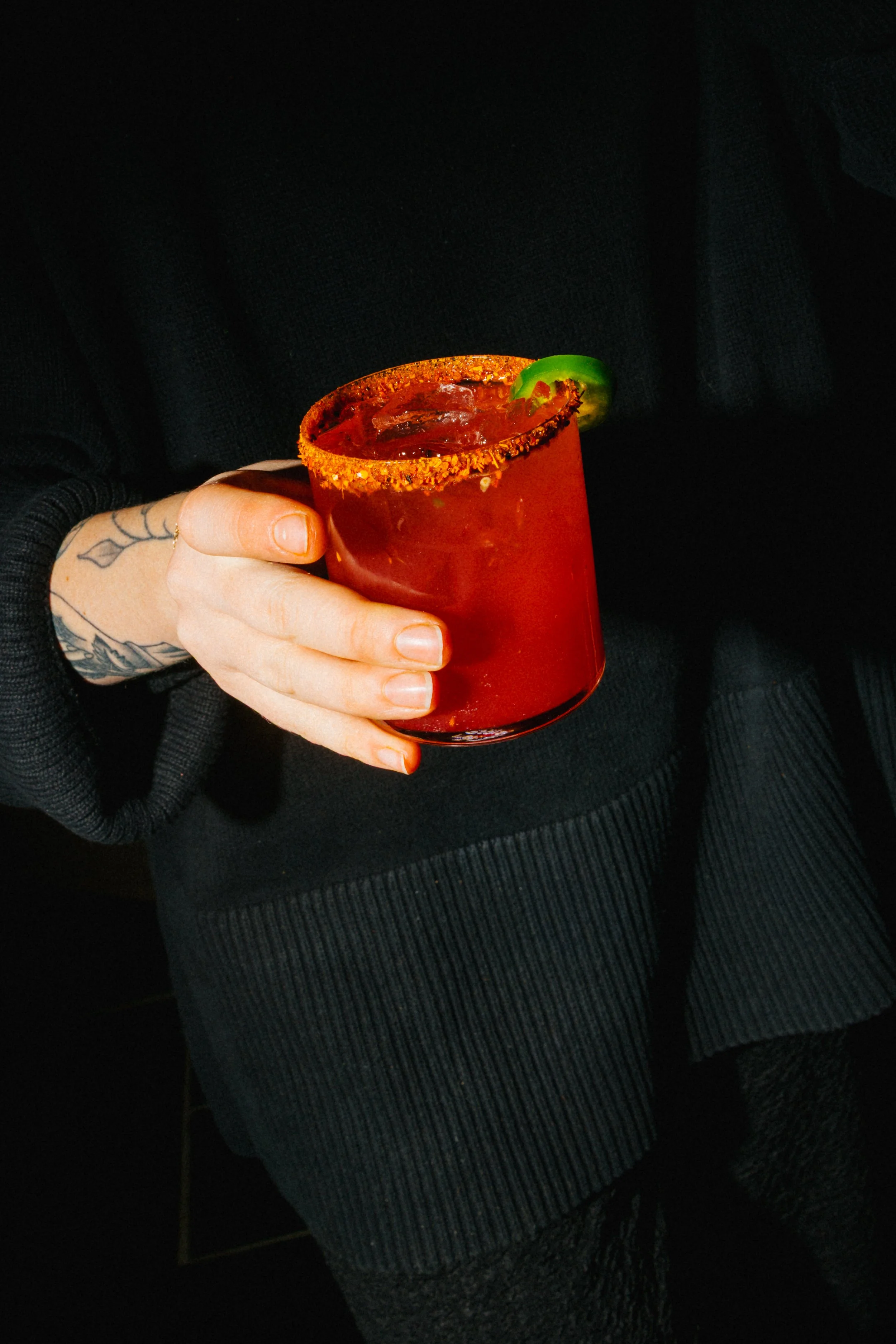 A person holding a red cocktail glass garnished with a slice of jalapeño, with the person's hand showing a tattoo of a floral design.