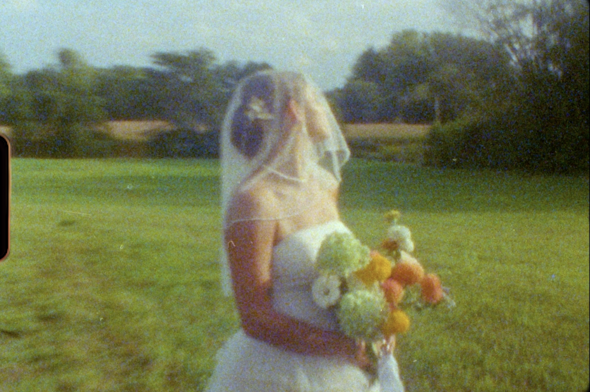 A woman in a wedding dress standing outdoors in a grassy field, holding a bouquet of flowers, with trees and a person in the background.