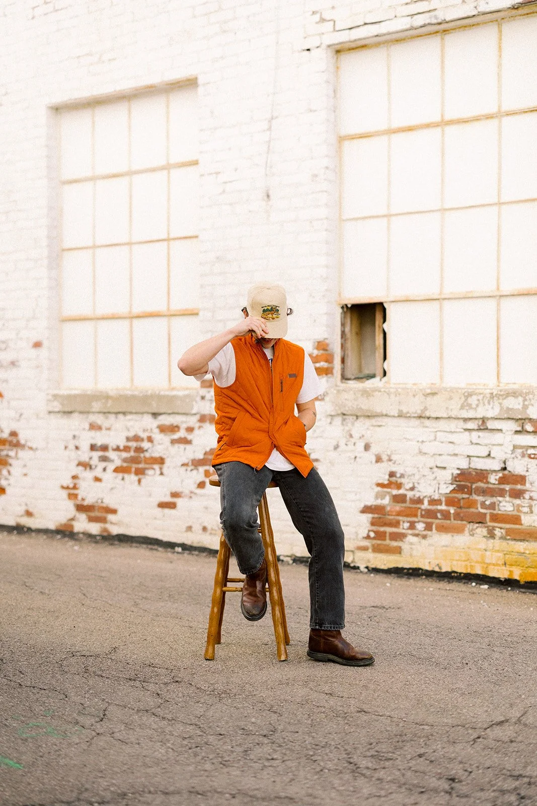 Person sitting on a wooden stool in front of a graffiti wall in an alleyway, wearing a beige cap, orange vest, white t-shirt, dark jeans, and brown boots, with hand resting on hat.
