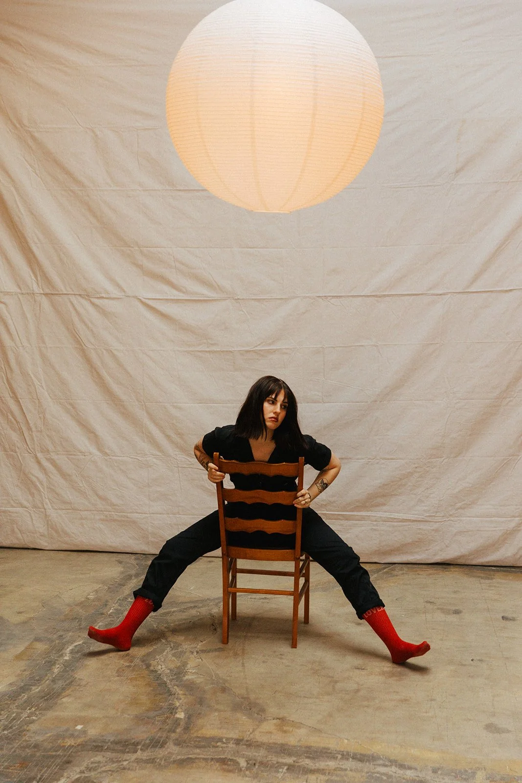 A woman with dark hair and tattoos poses sitting backward on a wooden chair with legs spread wide apart, wearing black clothing and red socks, in front of a beige backdrop and a hanging paper lantern.
