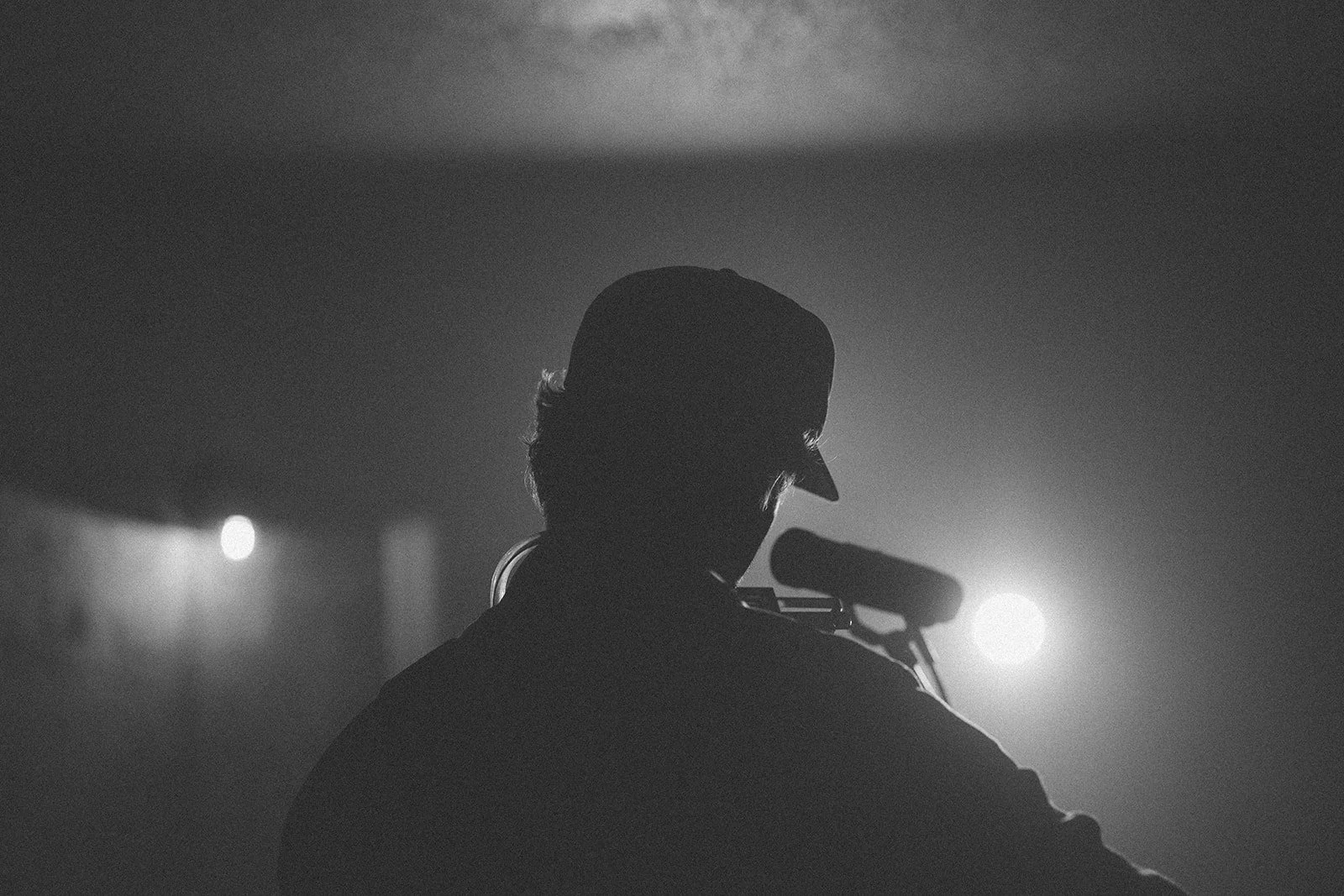 A black and white silhouette of a man with a cap, standing in front of a microphone, with bright stage lights behind him.