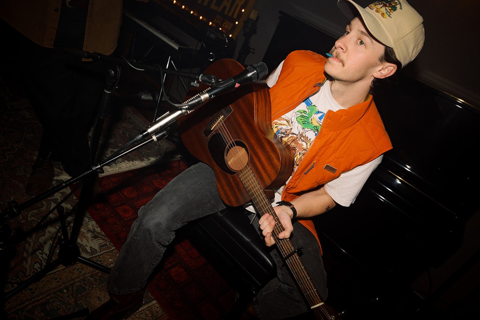 A man with a mustache wearing a beige cap, white t-shirt with colorful designs, and an orange vest, sitting on a black stool while holding an acoustic guitar and singing into a microphone on a stand in a dimly lit room.