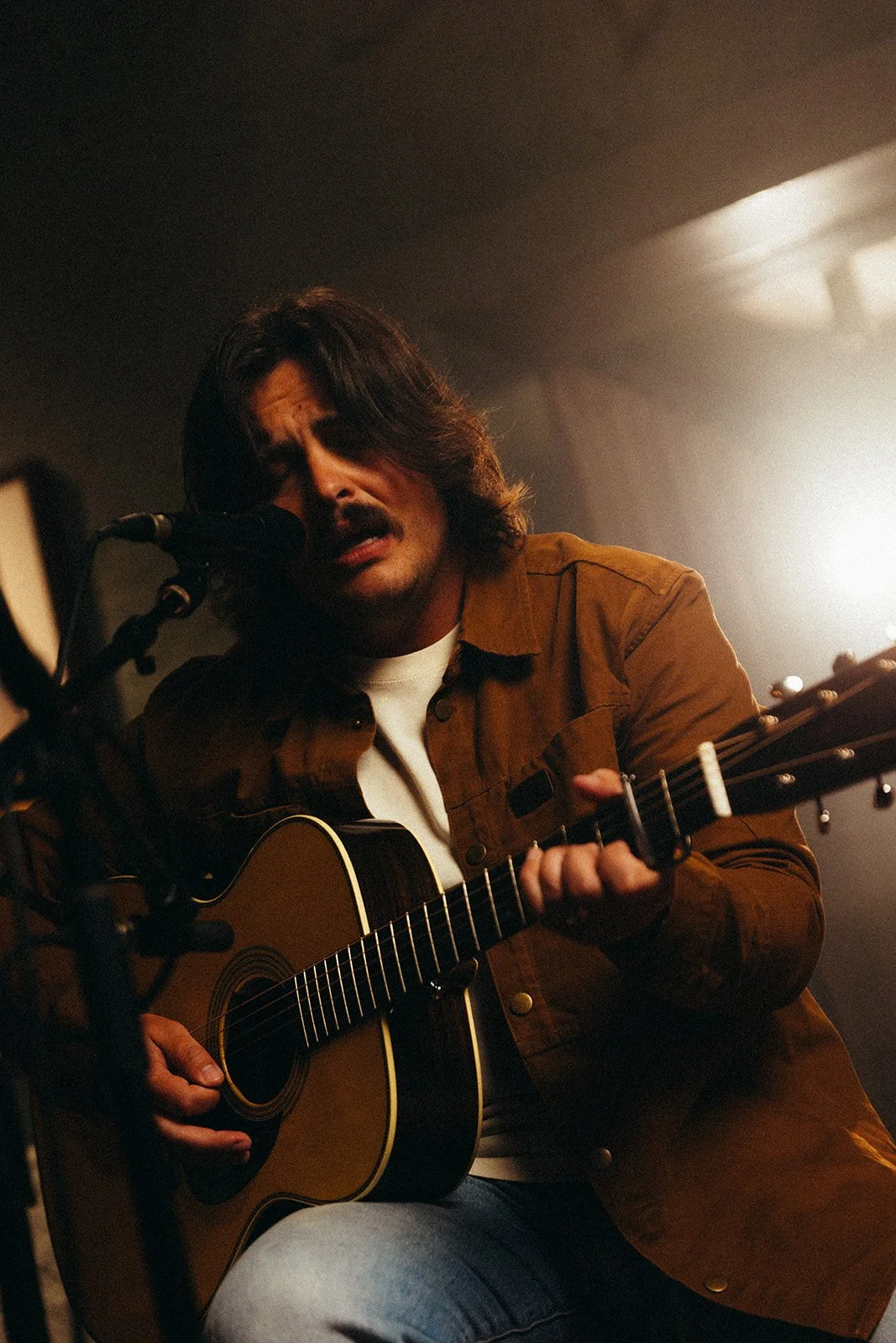 A man with long dark hair and a mustache singing and playing an acoustic guitar in a dimly lit room.