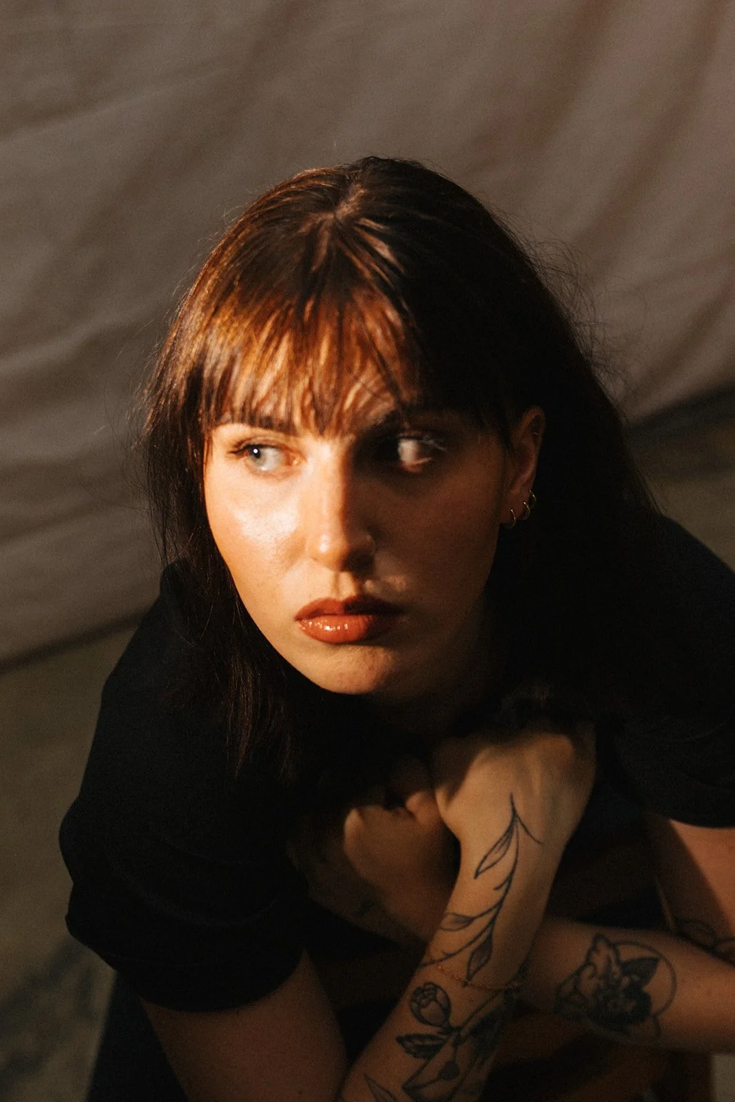 A woman with dark hair and tattoos on her arm, looking to her right with a serious expression, in a dimly lit environment.