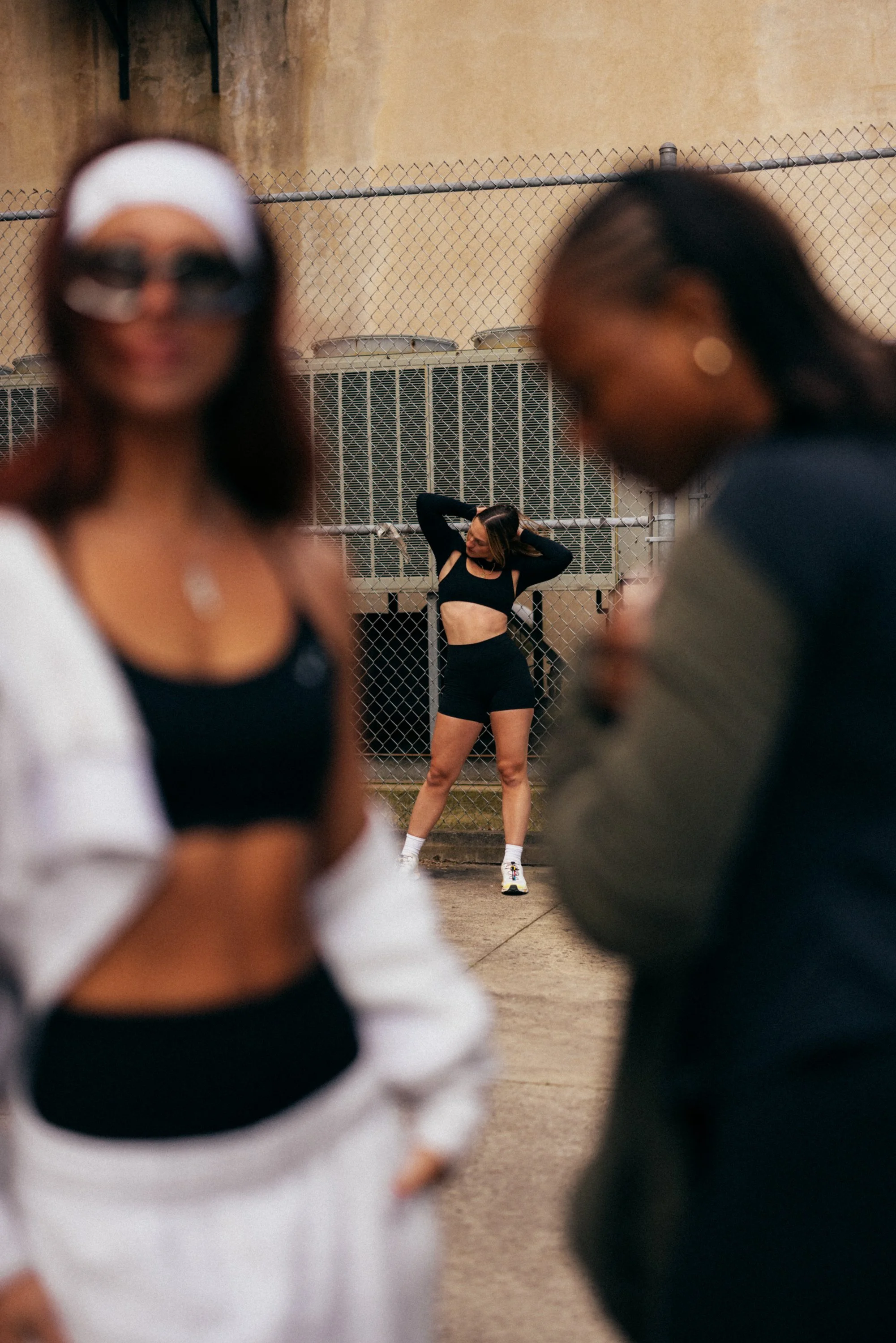 Blurred foreground of two women and a man with a woman in the background standing against a chain-link fence, wearing athletic clothes, with one hand behind her head, in an outdoor sports or gym setting.