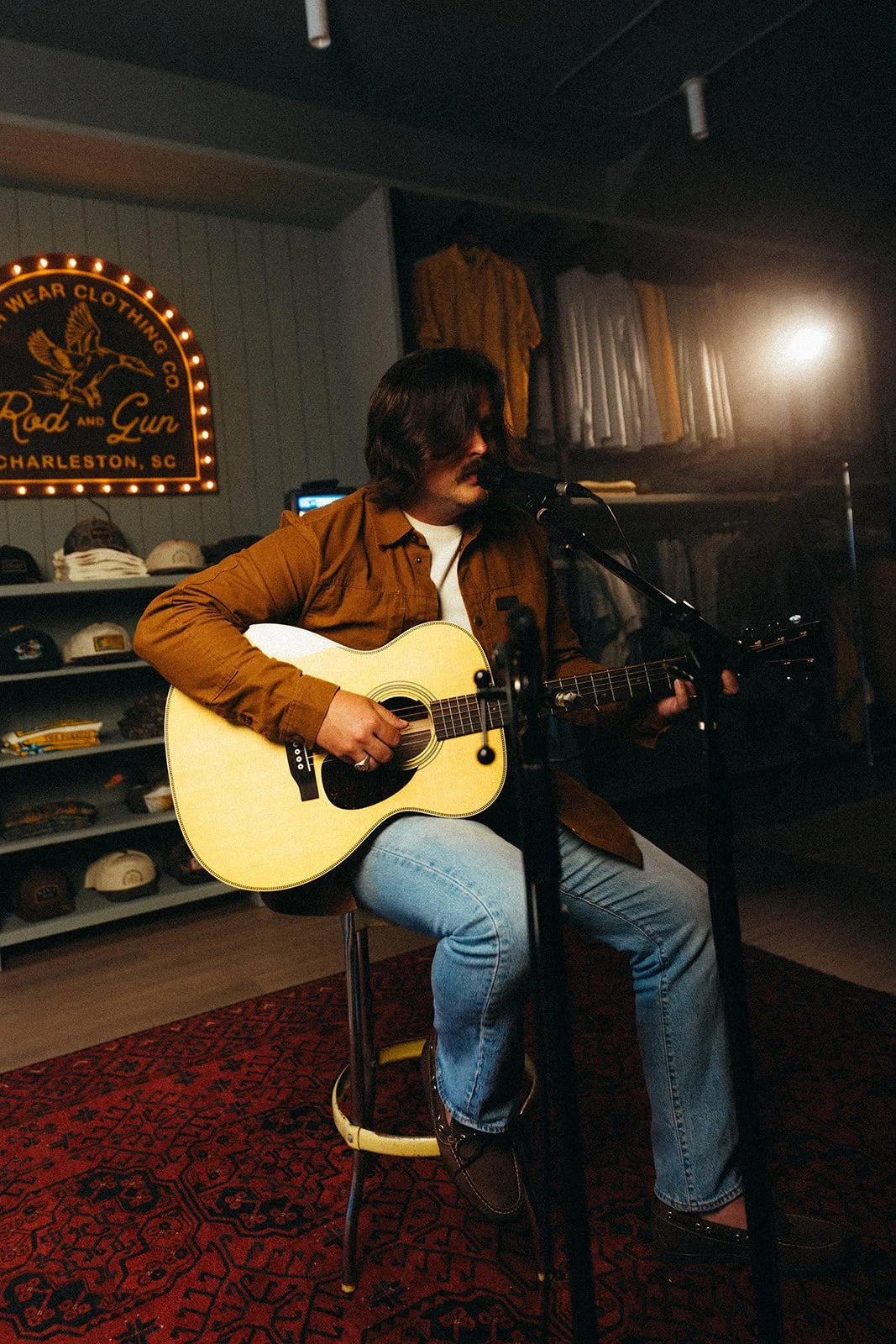 A man with shoulder-length dark hair and a mustache, wearing a brown jacket and light blue jeans, is sitting on a stool playing an acoustic guitar and singing into a microphone in a cozy music venue. Behind him, there are shelves with caps and a sign