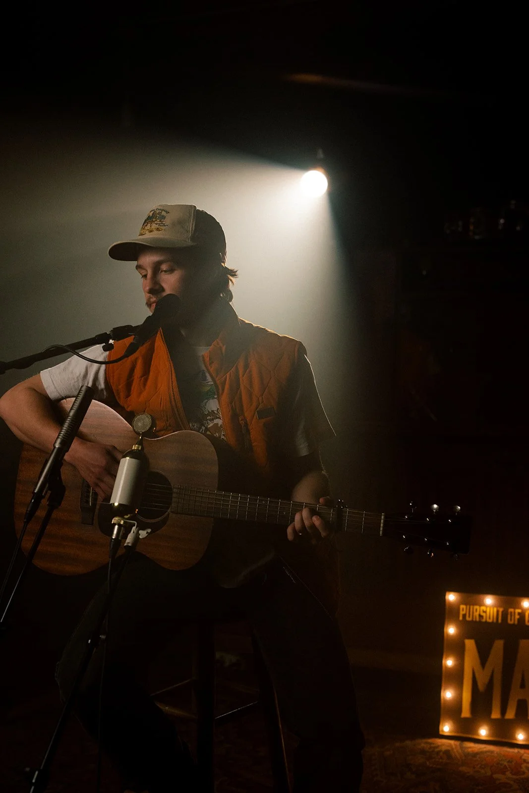 Person performing with a guitar on stage, wearing a cap and outside light shining behind.