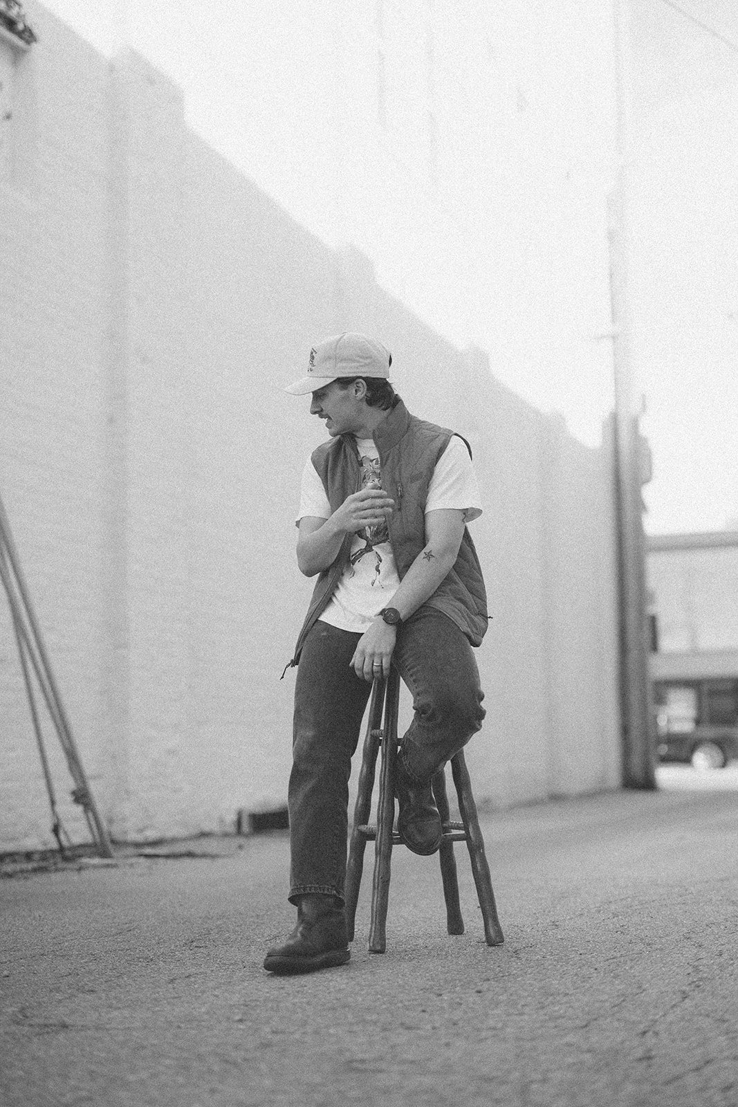 A young man sitting on a wooden stool in an alley, wearing a baseball cap, a vest, t-shirt, jeans, and boots, looking down at his hand.
