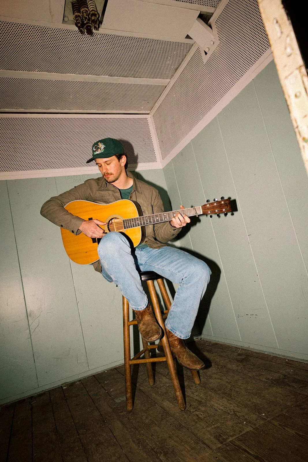 A young man wearing a green cap, brown jacket, blue jeans, and brown boots sitting on a wooden stool playing an acoustic guitar in a small room with light green walls and a wooden floor.