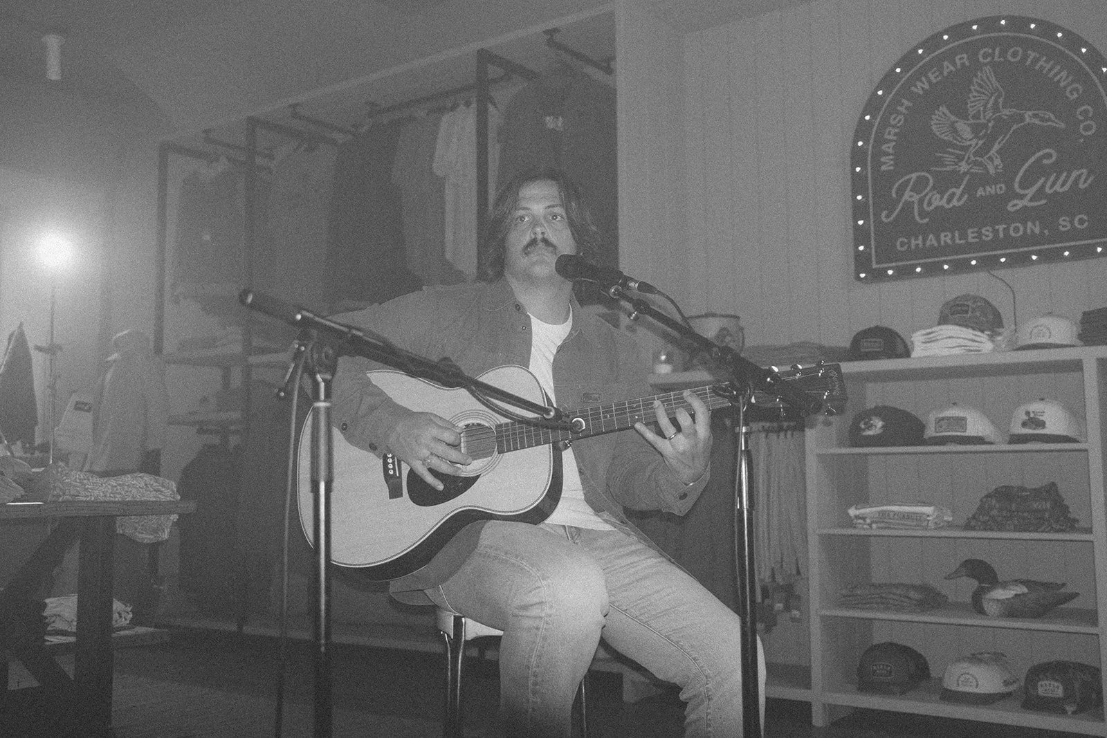 A man with long hair and a mustache is playing an acoustic guitar and singing into a microphone in a room with merchandise shelves and a sign that reads "Maret Wear Clothing Co. Rock and Guitars, Charleston, SC."