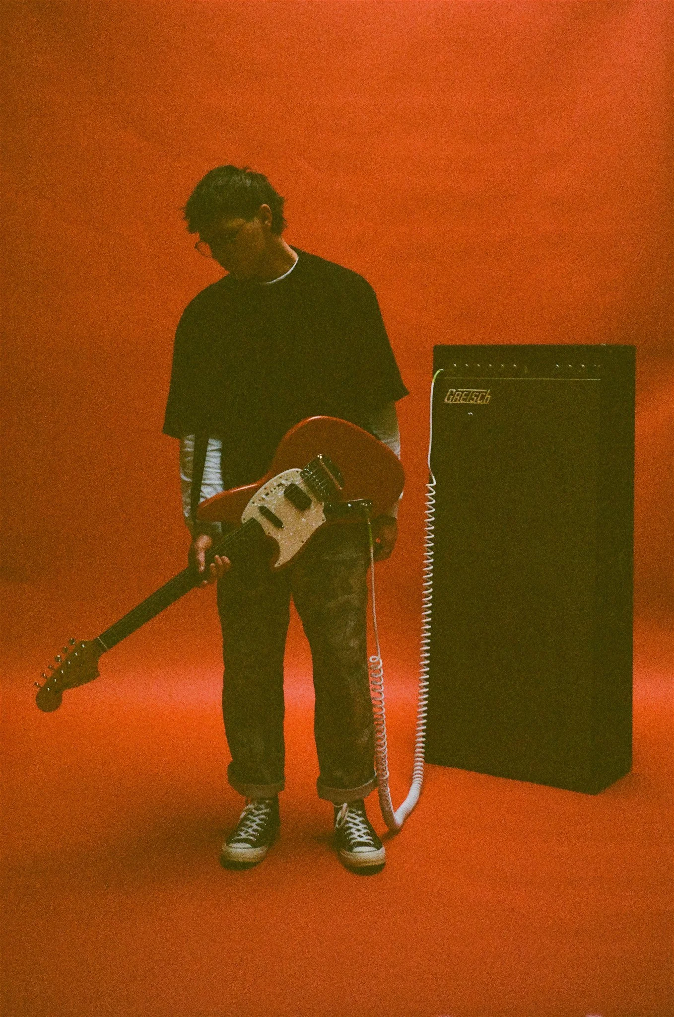 A person standing in a room with orange walls, holding an electric guitar, facing downward, next to an amplifier.