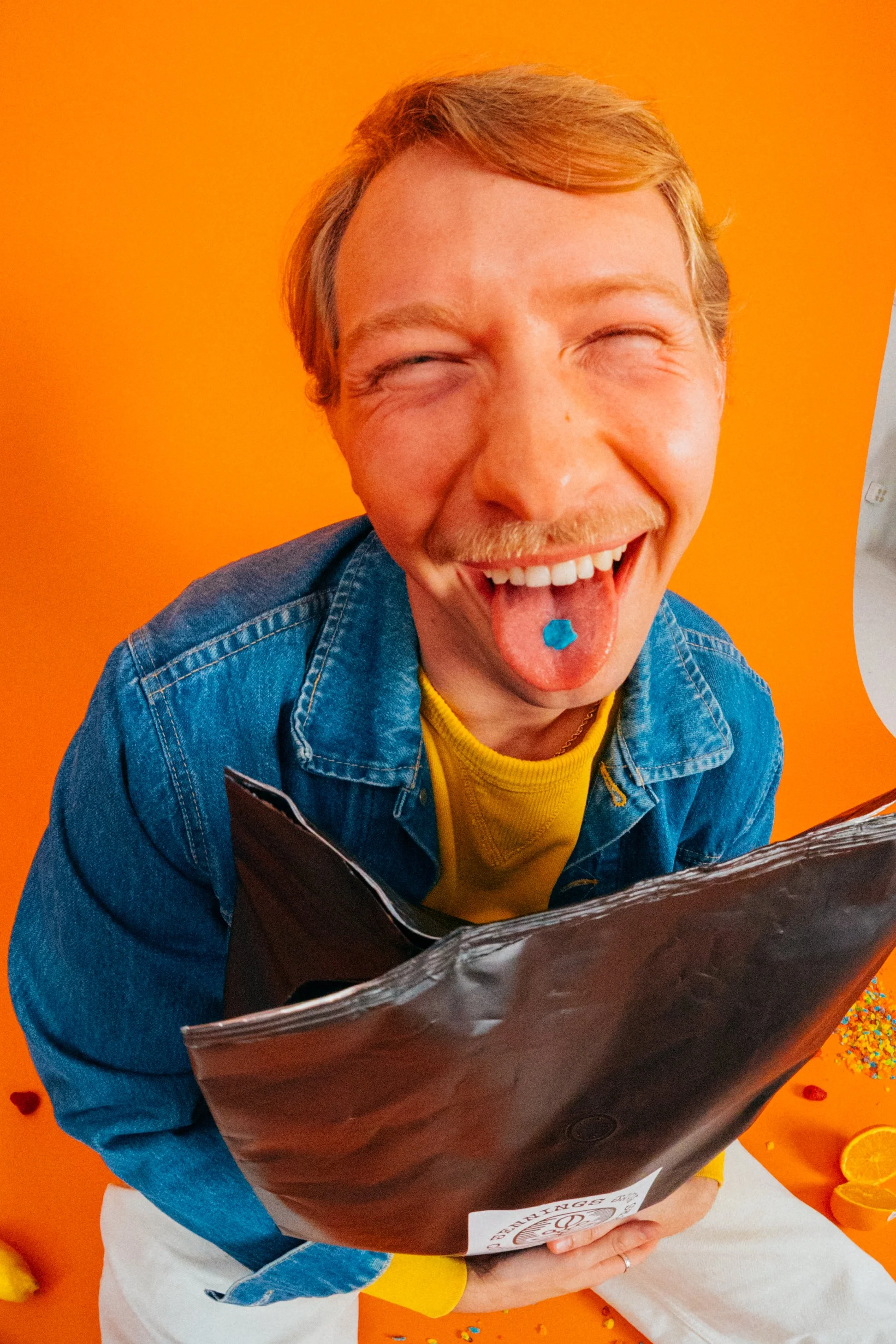 Young man with red hair, smiling and licking blue candy, wearing a denim jacket and yellow shirt, in front of an orange background with party decorations.