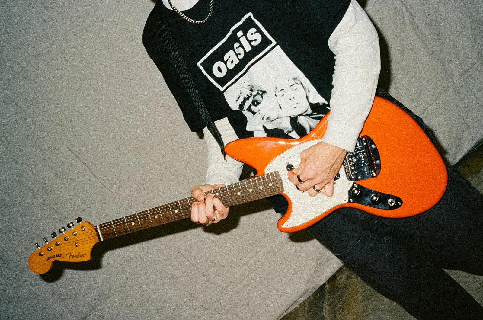 Person lying on the ground playing an orange electric guitar, wearing a black and white Oasis band t-shirt and silver rings.