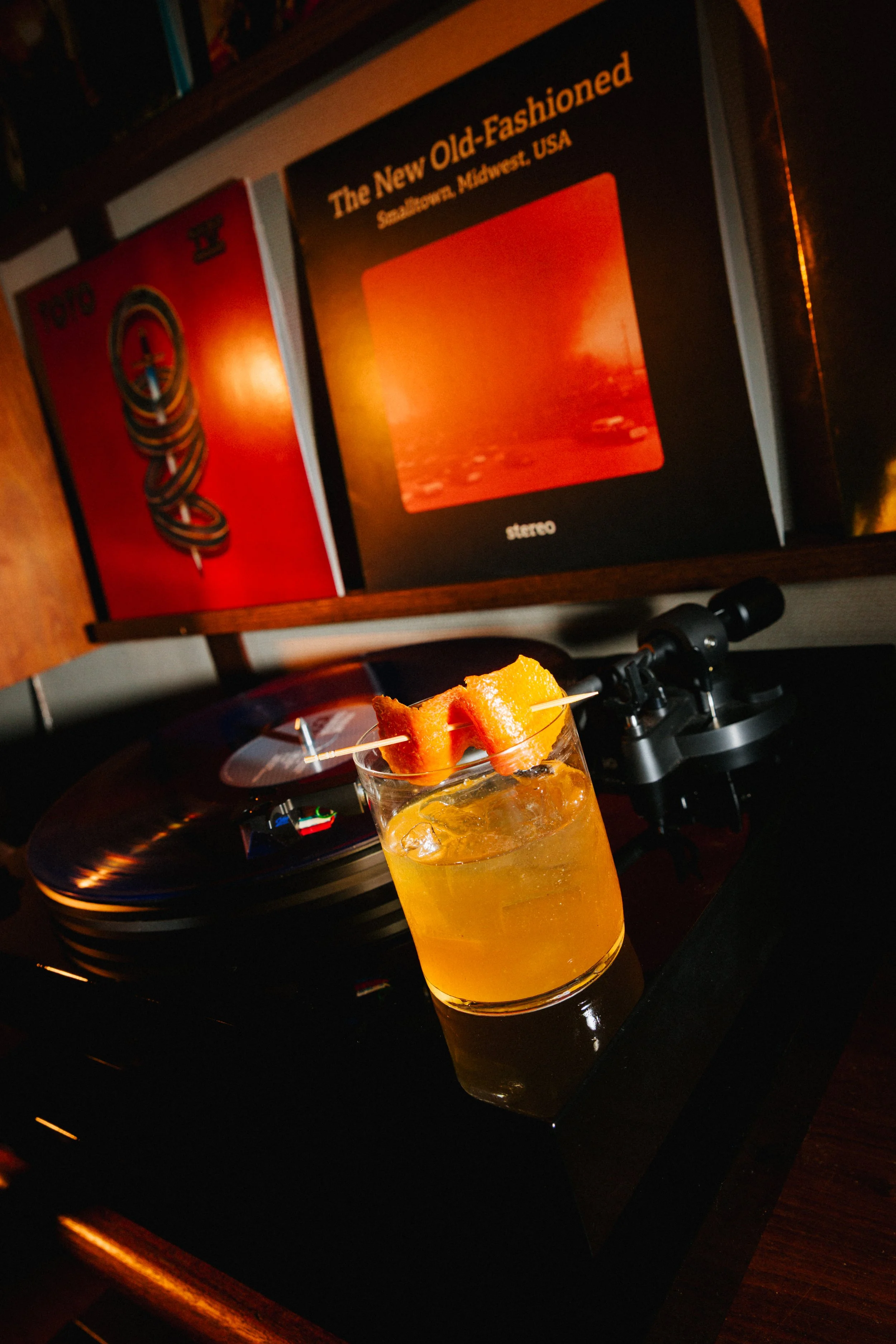 A glass of orange-colored cocktail garnished with a pieces of bread on a skewer, on a turntable with album covers in the background.