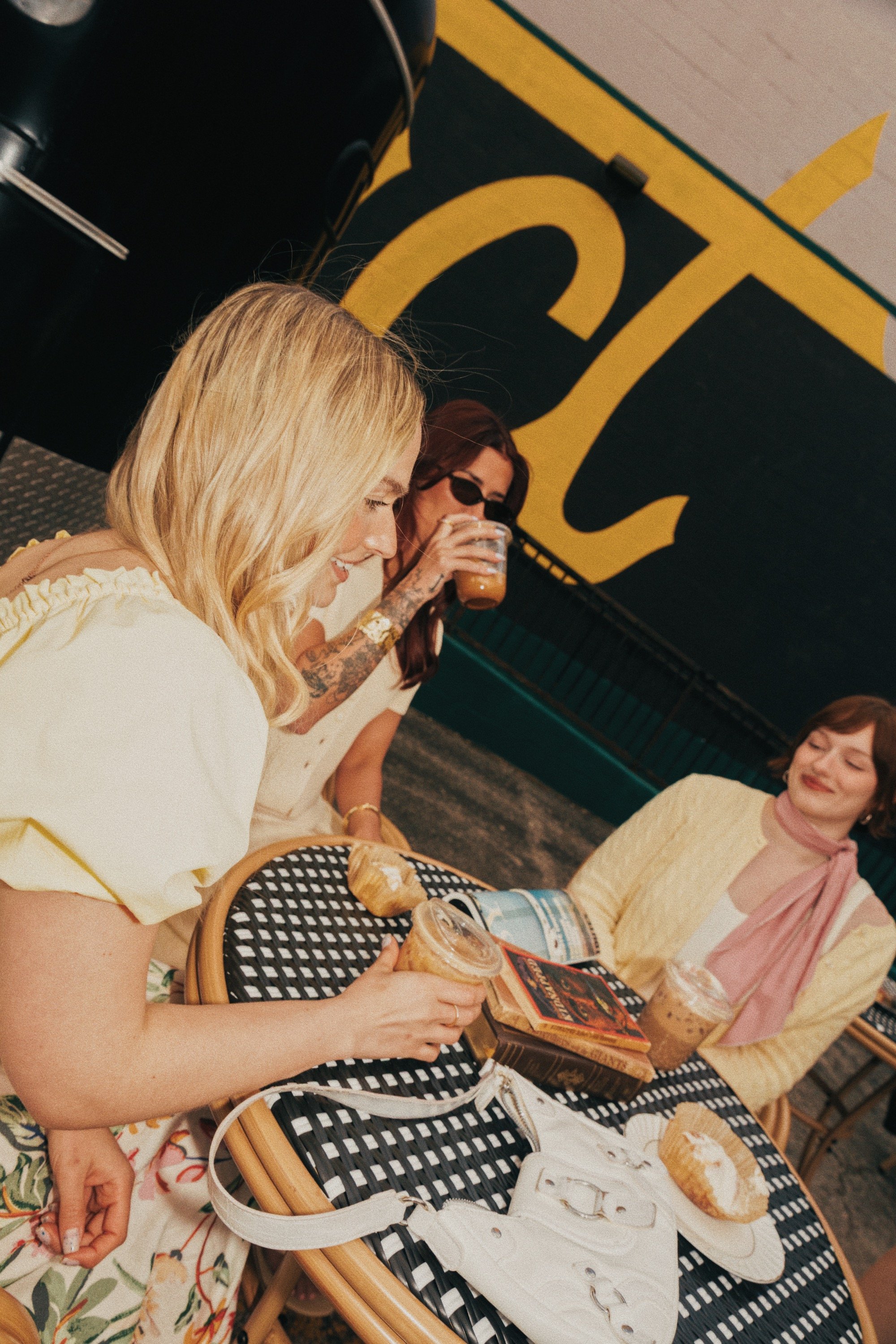 Several women sitting around a black and white checkered outdoor table, enjoying ice cream and snacks. One woman with blonde hair in a yellow shirt is in the foreground, and another woman with reddish hair, sunglasses, and tattoos is drinking iced co