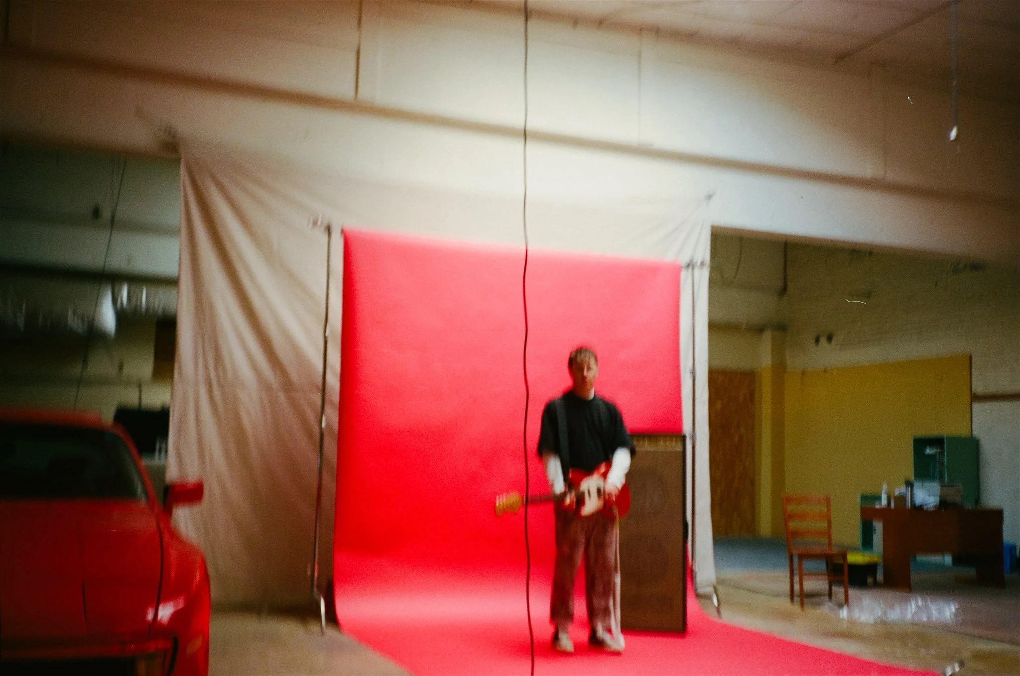 A person standing in front of a red backdrop in an indoor setting, holding a guitar with a car parked on the left side and a desk with a chair and office supplies on the right side.