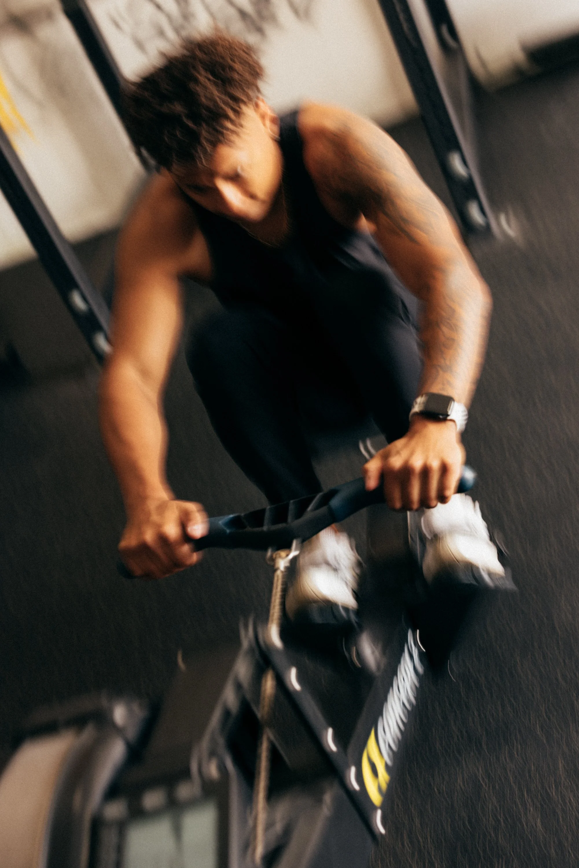 A person with short reddish-brown hair, wearing a black tank top and white sneakers, is seen from above riding a black skateboard with yellow branding on a gym or indoor Sports Hall with black flooring.