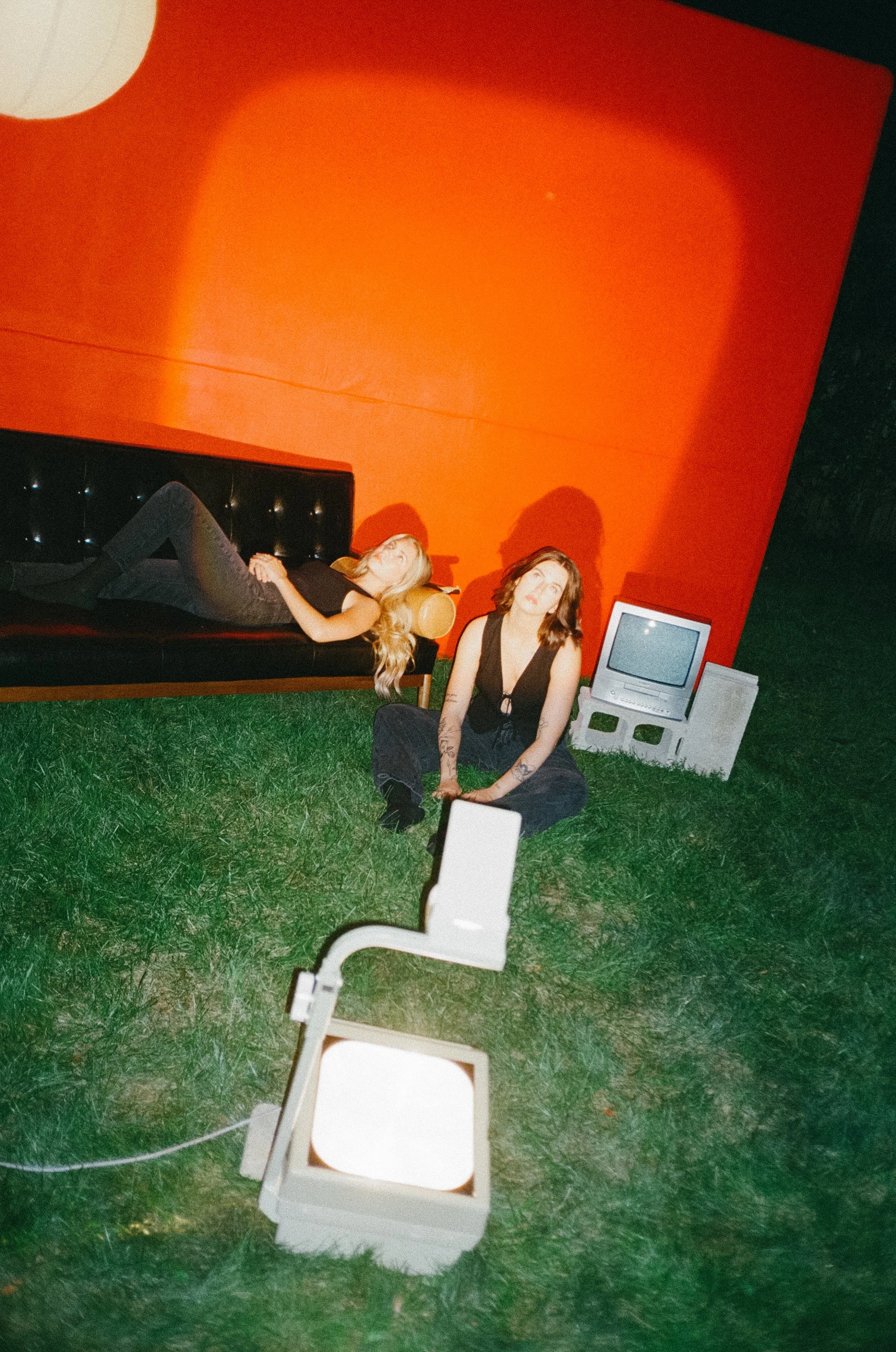 Two women with tattoos in black clothing, one lying on a black couch and one sitting on green grass, surrounded by vintage electronics including a CRT monitor and a projector, in front of an orange wall at night.