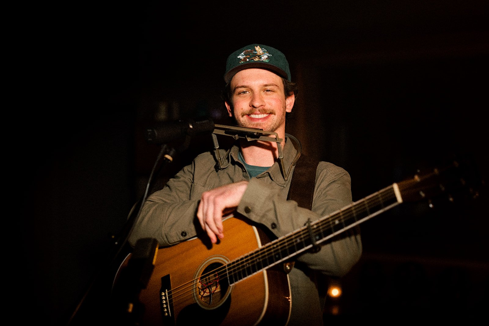A man wearing a green baseball cap with a bird emblem, smiling, with an acoustic guitar and a harmonica holder around his neck, performing on stage in a dark setting.