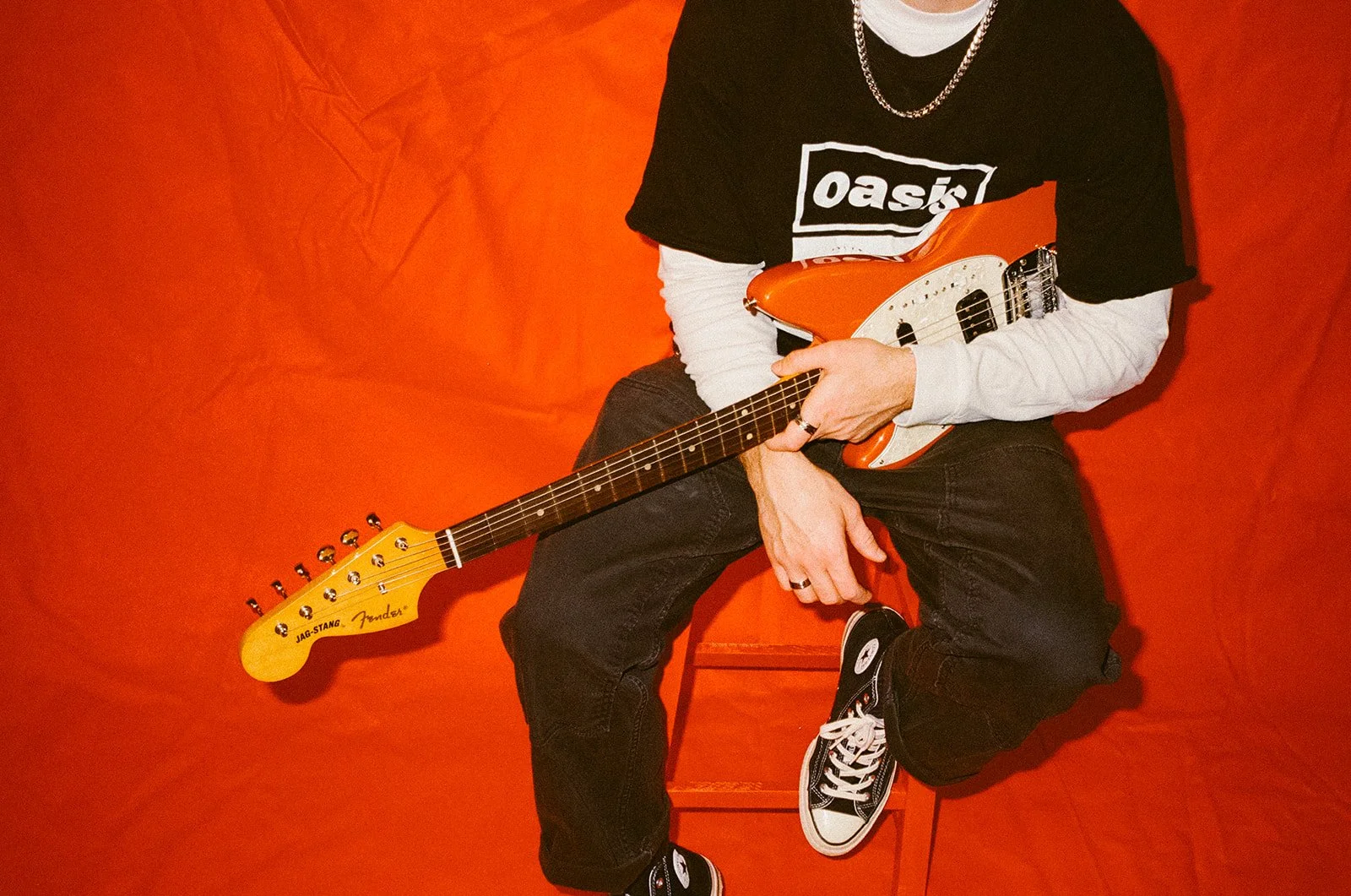 A person wearing black sneakers, black jeans, and layered clothing holds an orange electric guitar while sitting on a small orange table against an orange background.