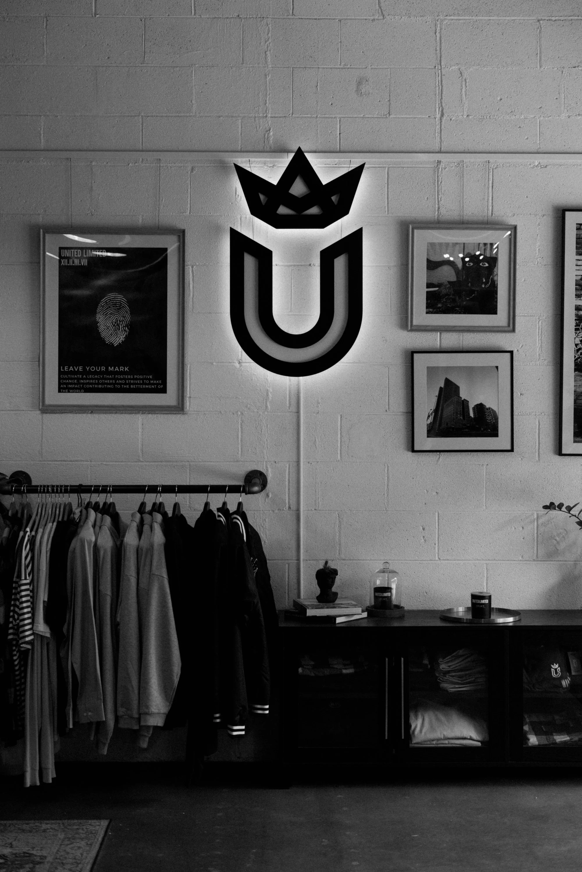 Interior of a clothing store with a wall display featuring framed artwork and a wall-mounted logo of a crown and U shape. Racks of clothes are below the display, with folded clothes and decorative items on a table.