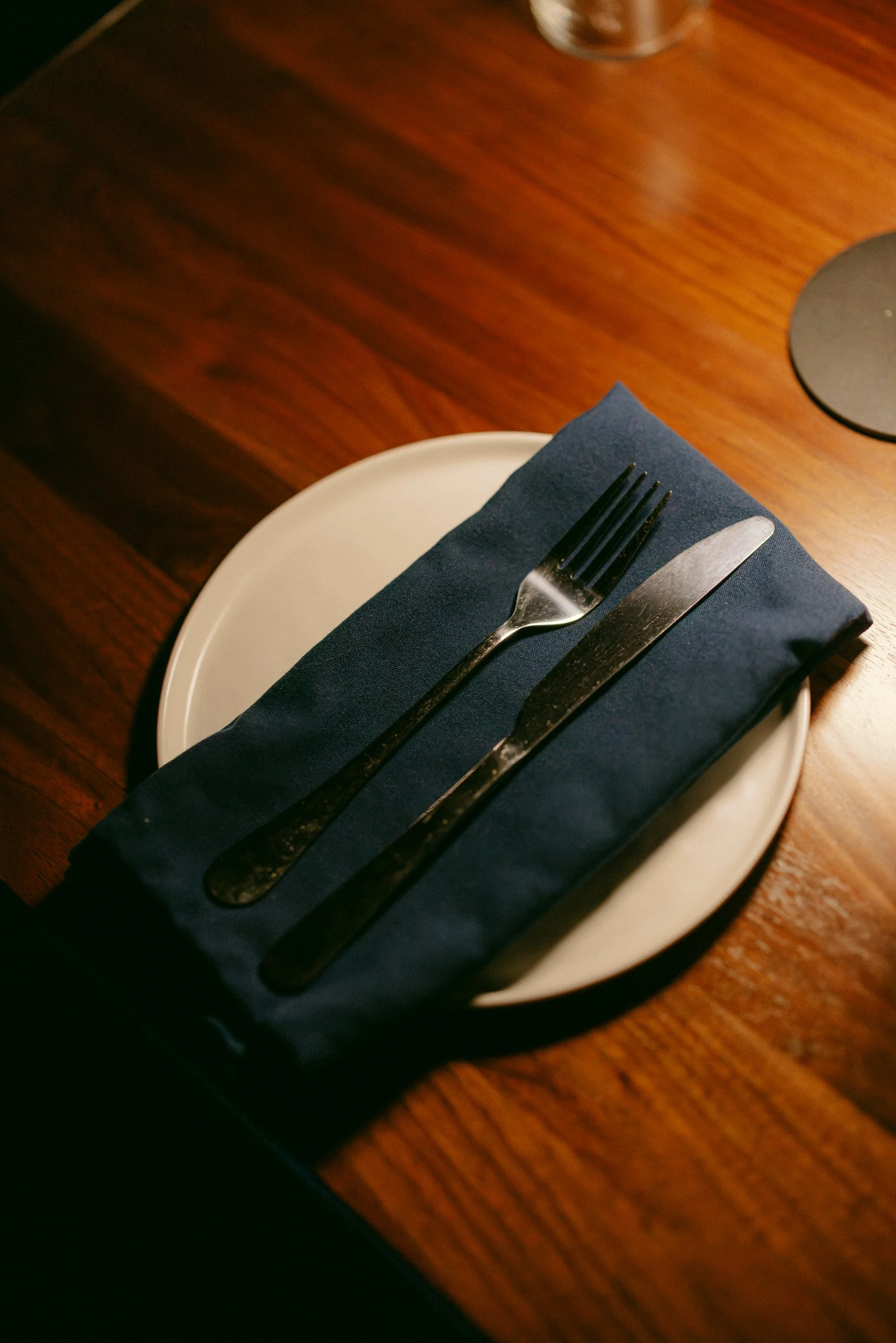A white plate with a folded dark blue napkin, and a fork and knife resting on top, placed on a wooden table.