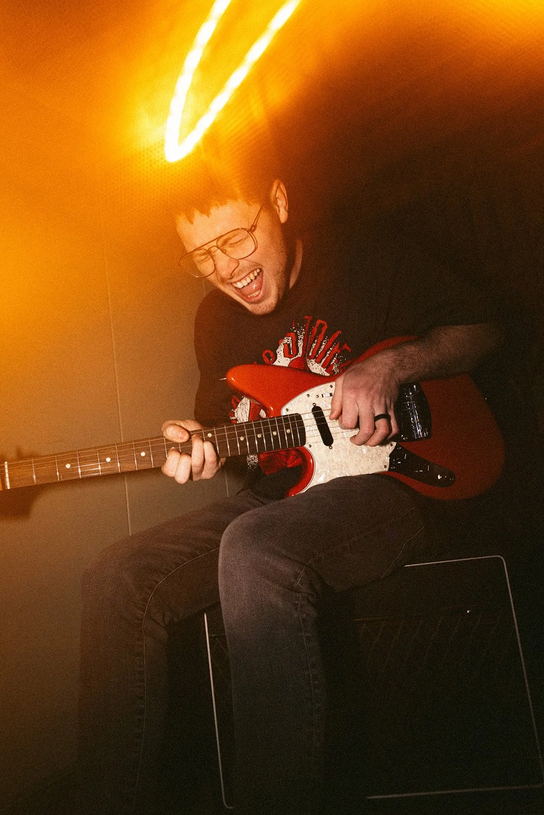A young man with glasses and dark hair, wearing a black T-shirt, is sitting on a black stool playing an electric guitar and smiling joyfully. The photo is taken in a setting with warm, yellow-orange lighting.